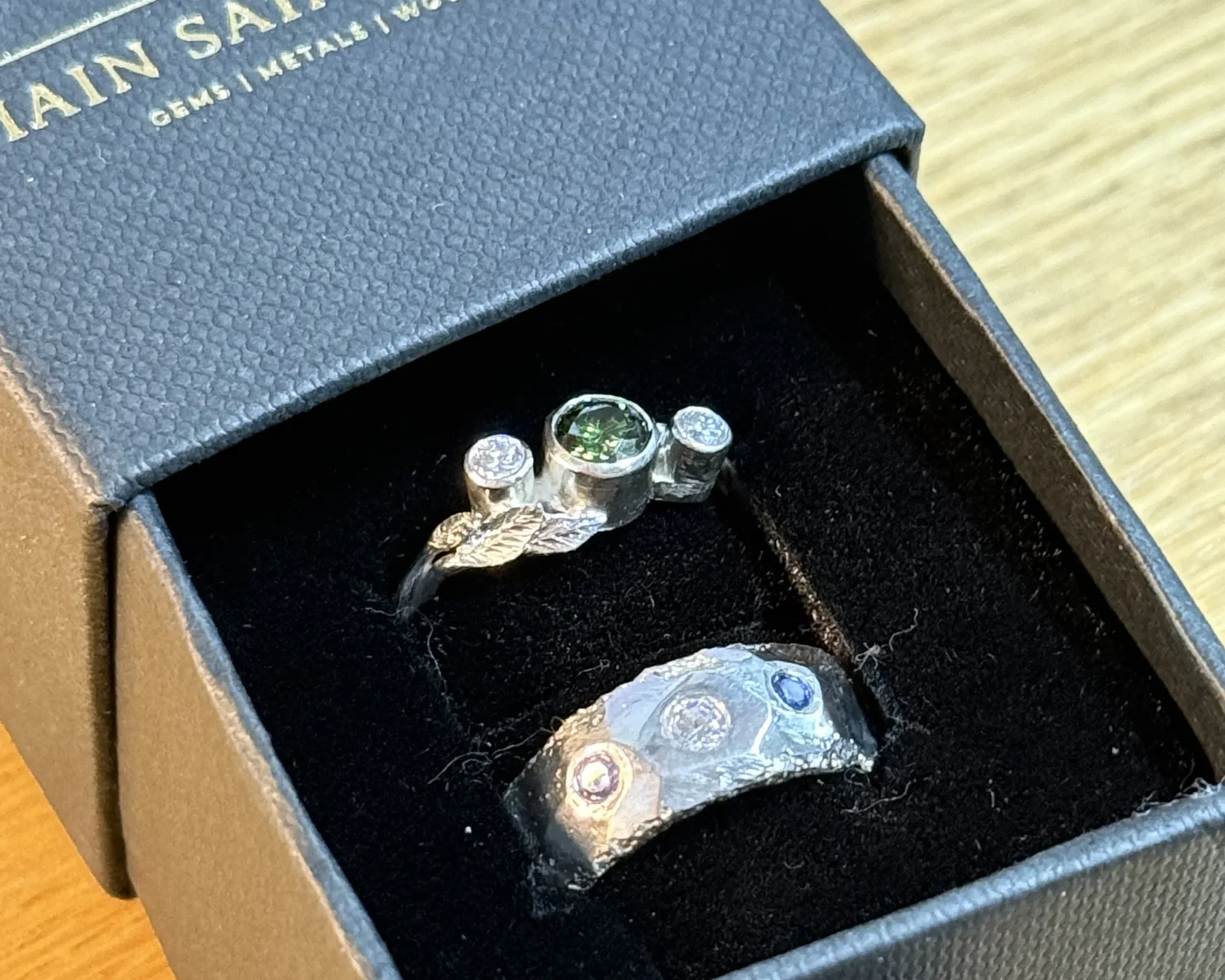 A jewelry box with a textured surface containing a silver ring and matching earrings with circular glittering stones, with a green gemstone in the ring.