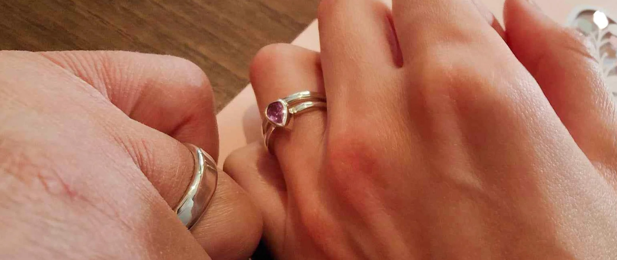Close-up of two hands wearing matching wedding rings, one with a pink sapphire. The hands are resting on a wooden surface.