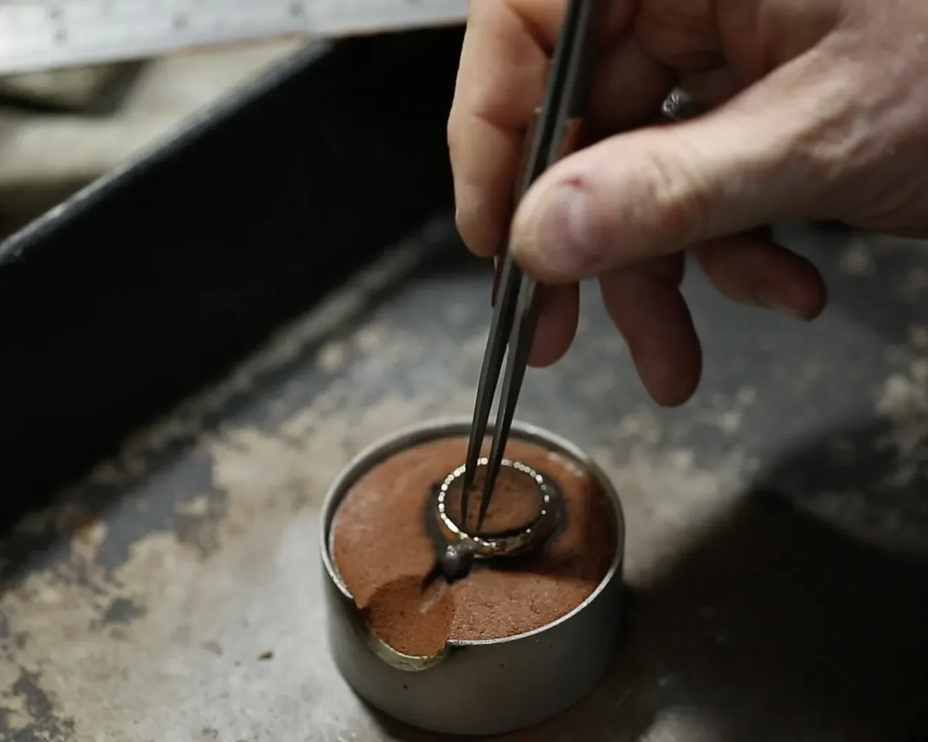 A person using tweezers to manipulate a small ring embedded in a circular object filled with brown powder, possibly a gemstone or mineral sample, on a dark work surface.