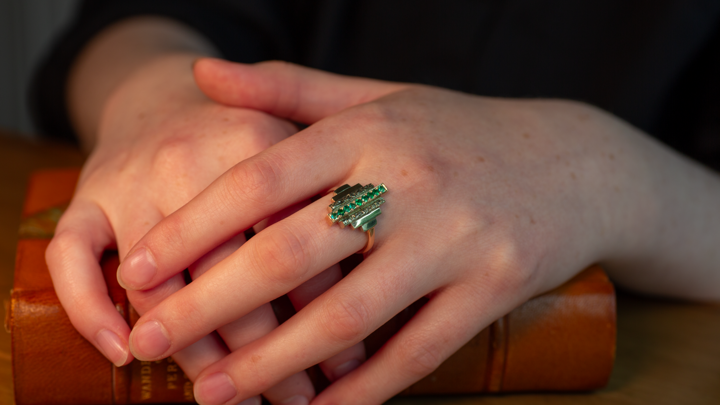 Close-up of a woman's hands resting on a vintage book, wearing a art deco style ring with nine emeralds.