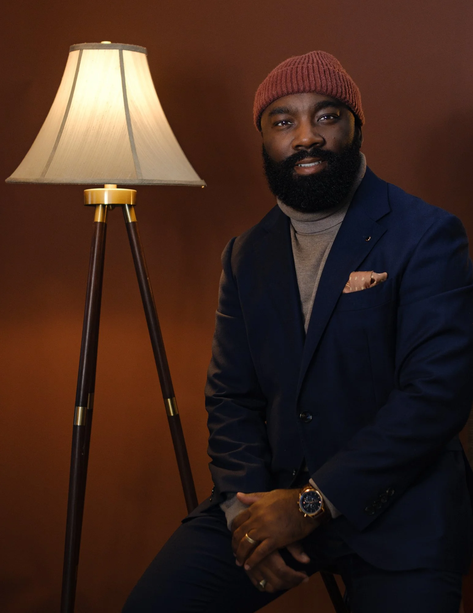 A man with a beard wearing a red beanie, navy blazer, gray turtleneck, and a watch, sitting near a standing lamp with a beige lampshade against a brown background.