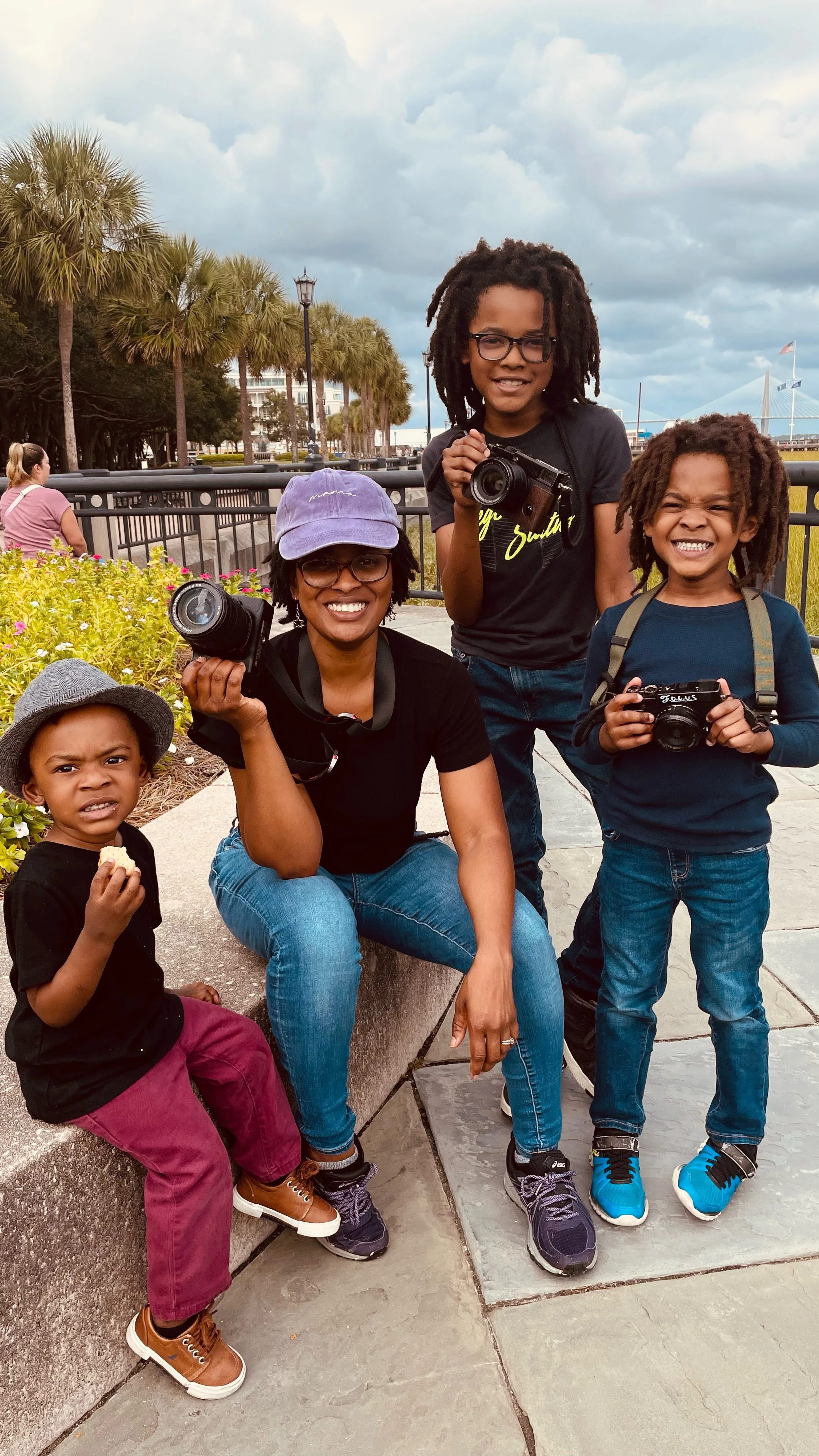 Group of five people, including three children and two women, outdoors with palm trees and cloudy sky, some holding cameras.