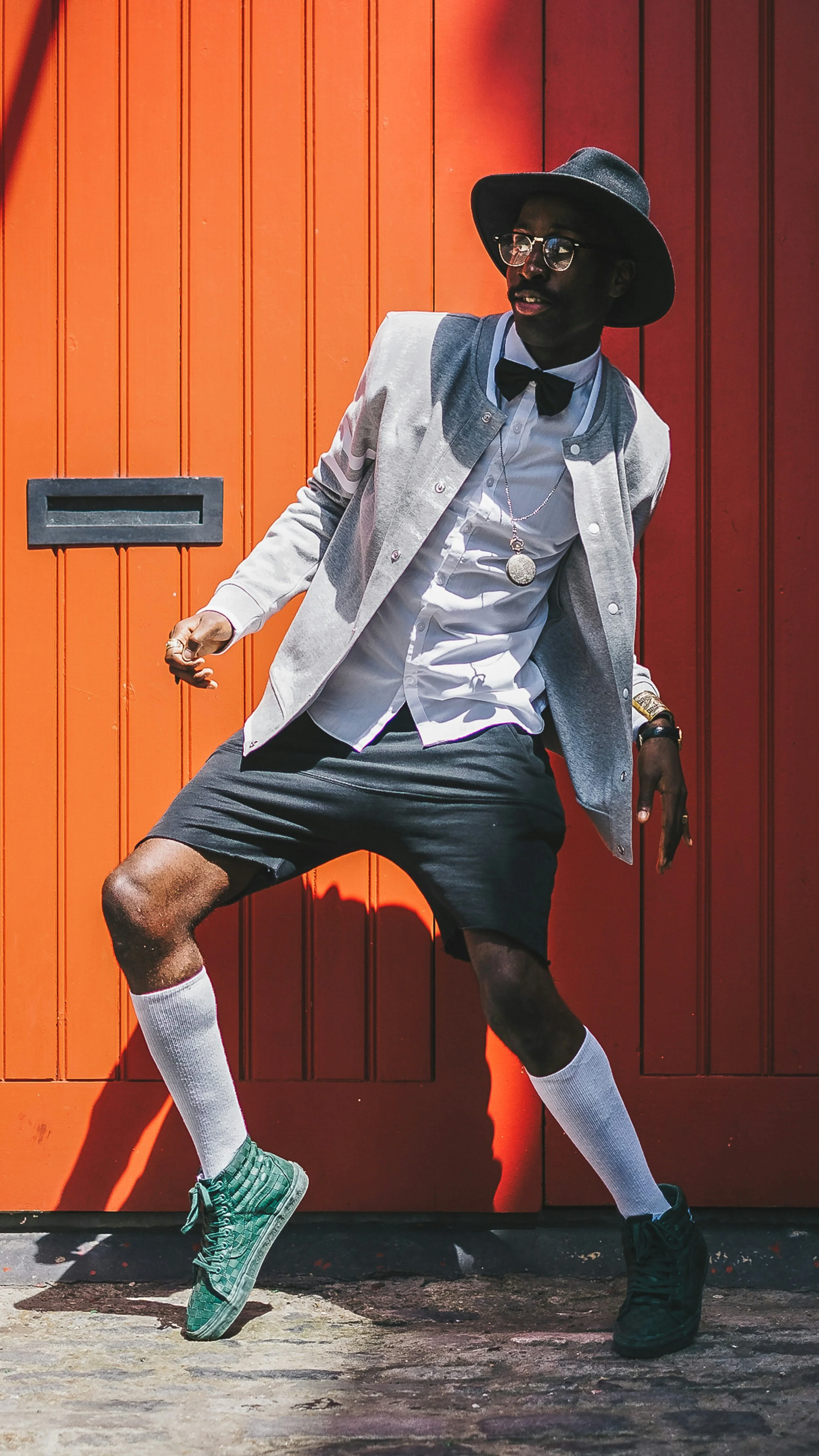 Man wearing a gray jacket, white shirt, black shorts, white socks, green sneakers, black hat, glasses, and bow tie standing against an orange wooden wall, striking a dynamic pose.