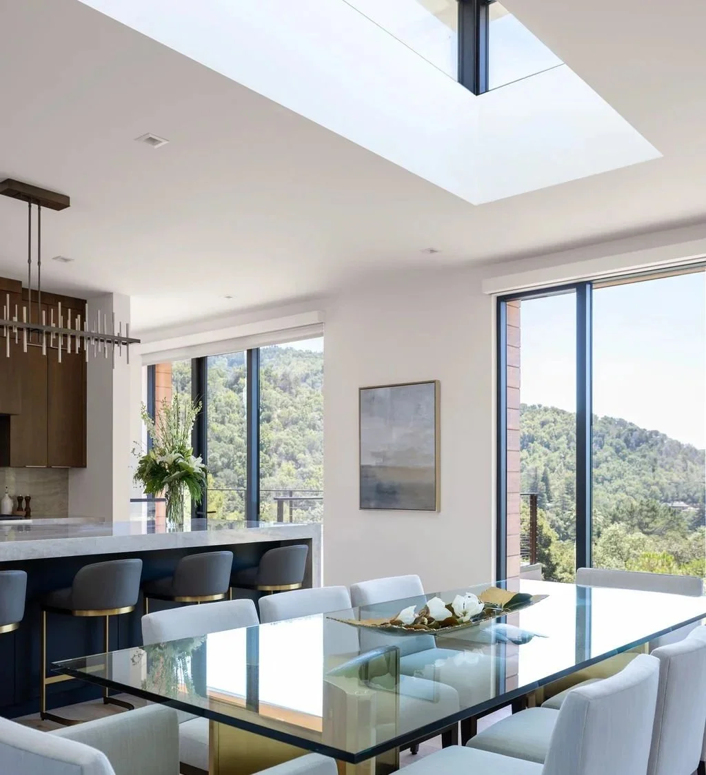 Modern dining area with glass table, white chairs, and a decorative centerpiece. Large windows provide a view of a forested landscape, and there's a kitchen island with bar stools in the background.