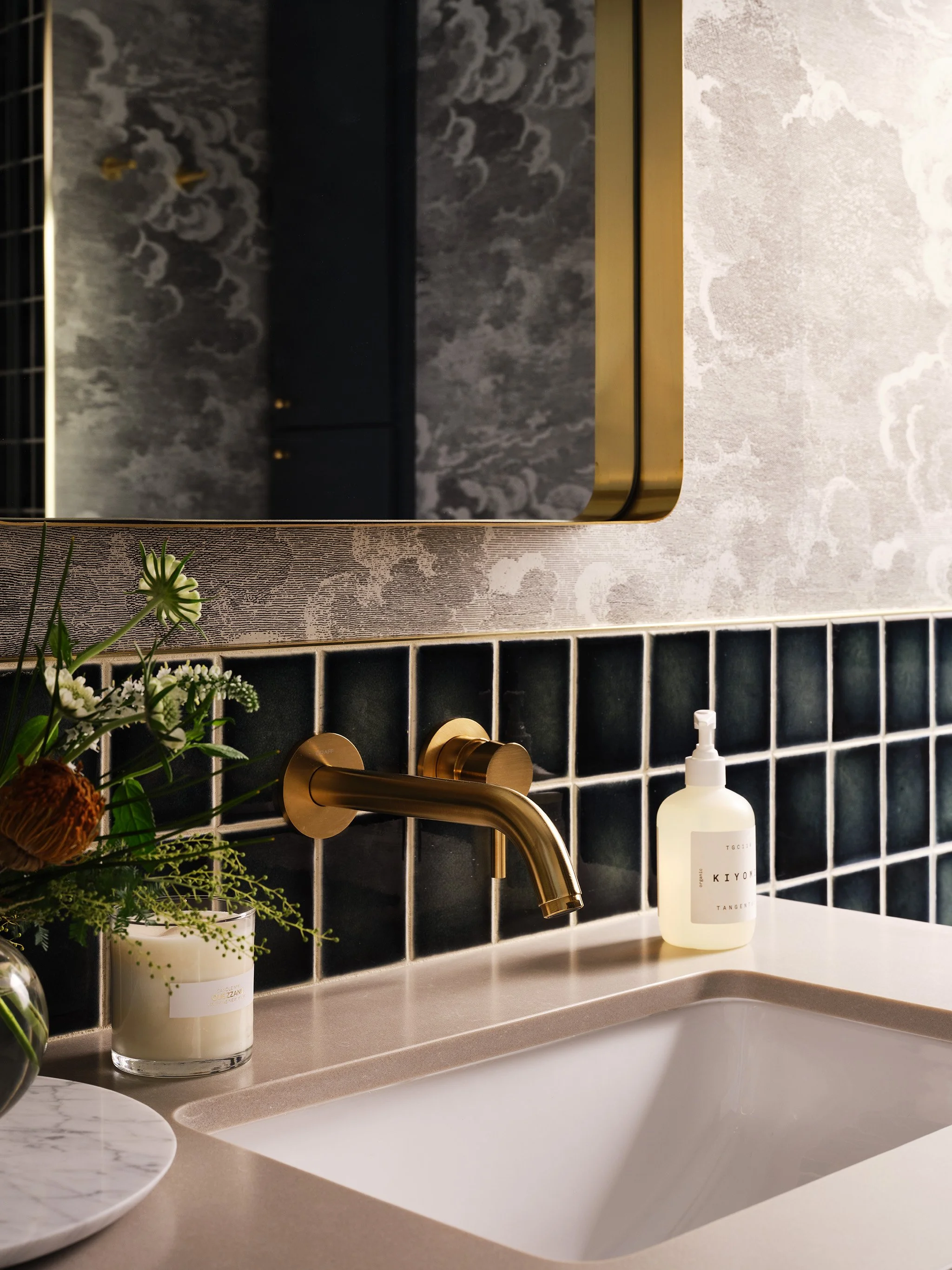 Modern bathroom sink with gold faucet, dark tiled backsplash, decorative plant, candle, and liquid soap dispenser.