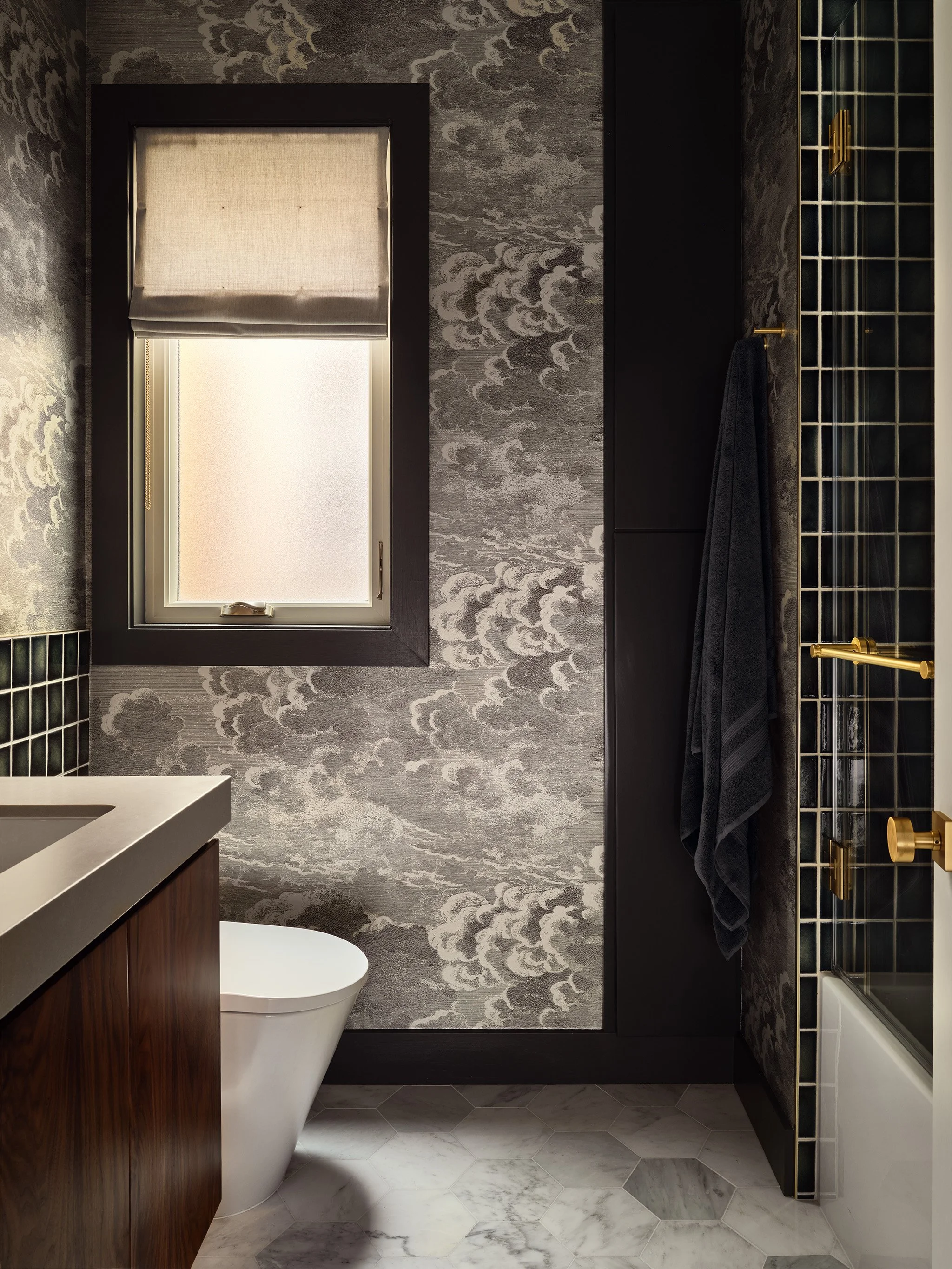 Modern bathroom with gray cloud-patterned wallpaper, a window with a linen roman shade, a white toilet, dark walnut vanity, hexagonal marble floor tiles, and a shower with brass fixtures.