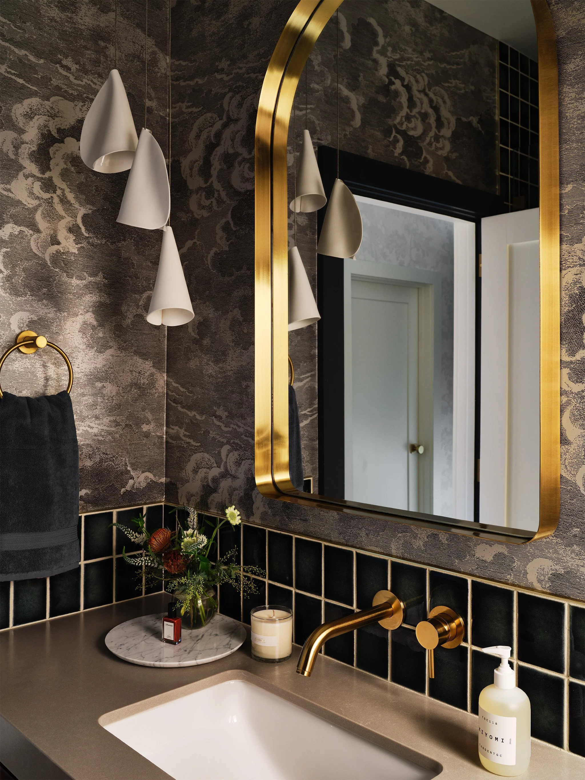 Modern bathroom interior with gold fixtures, a large mirror with gold frame, white ceramic pendant lights, a countertop with a sink, a black towel, a round marble tray with flowers, a candle, and hand soap. The walls have dark, cloud-patterned wallpa