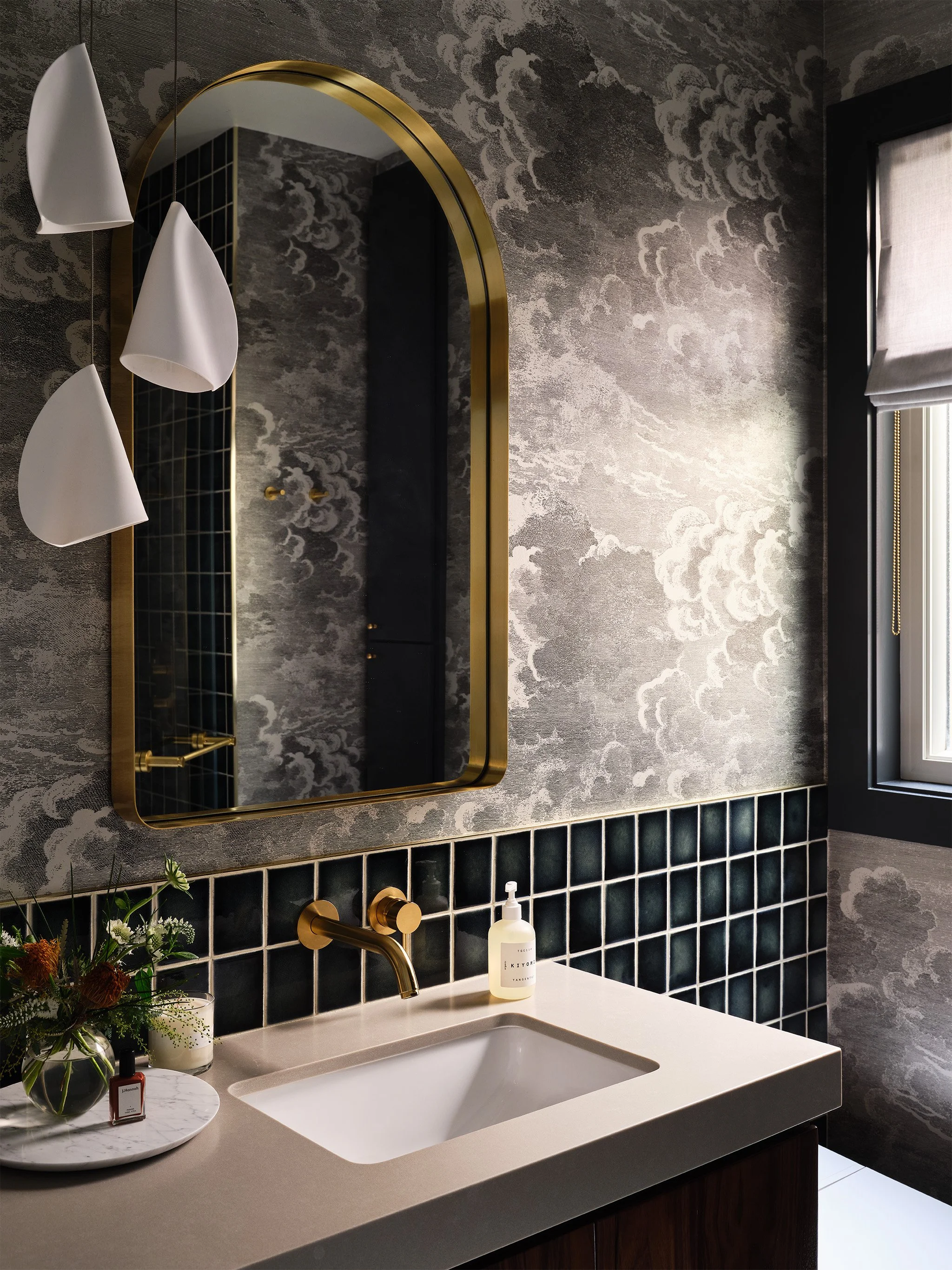 Modern bathroom interior with brass-framed mirror, patterned wallpaper, geometric lighting, tiled backsplash, white sink, and countertop with decorative items.