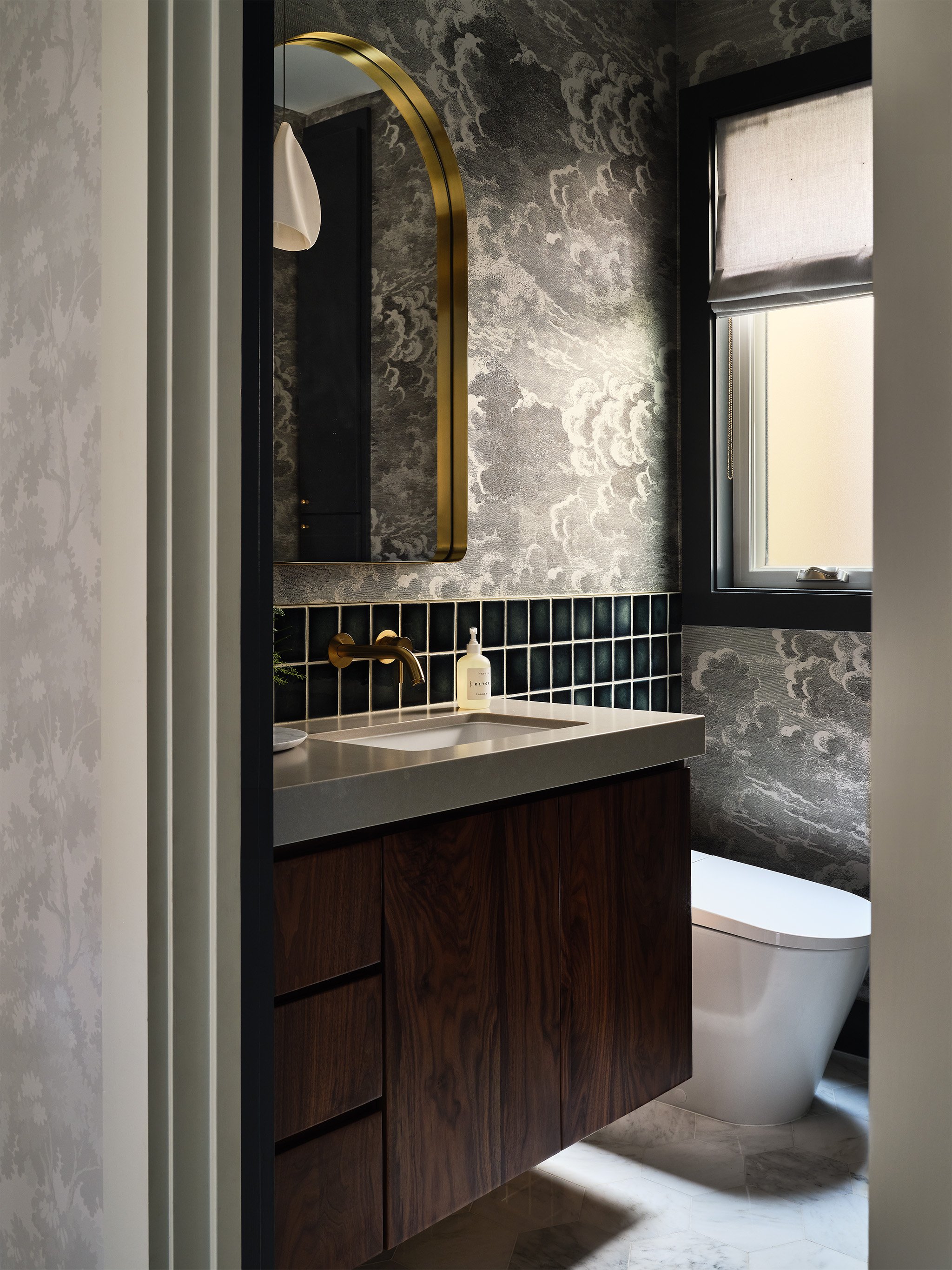 Modern bathroom with dark wood vanity, gold arched mirror, textured wallpaper, tiled backsplash, and sleek toilet.