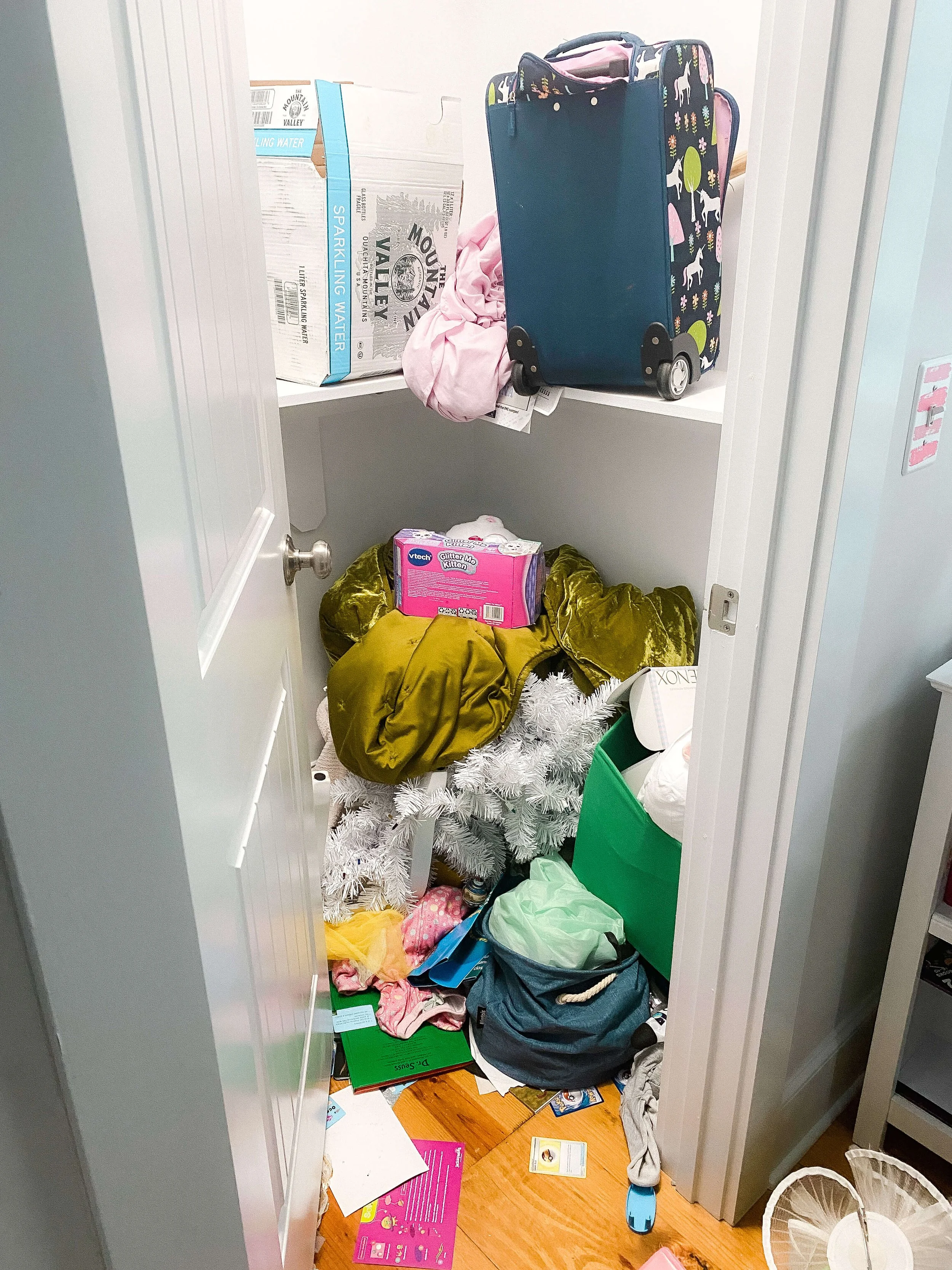  A small kids' closet before organizing, with a suitcase, cardboard boxes, a bagged holiday tree, and clothing and papers piled on the floor 
