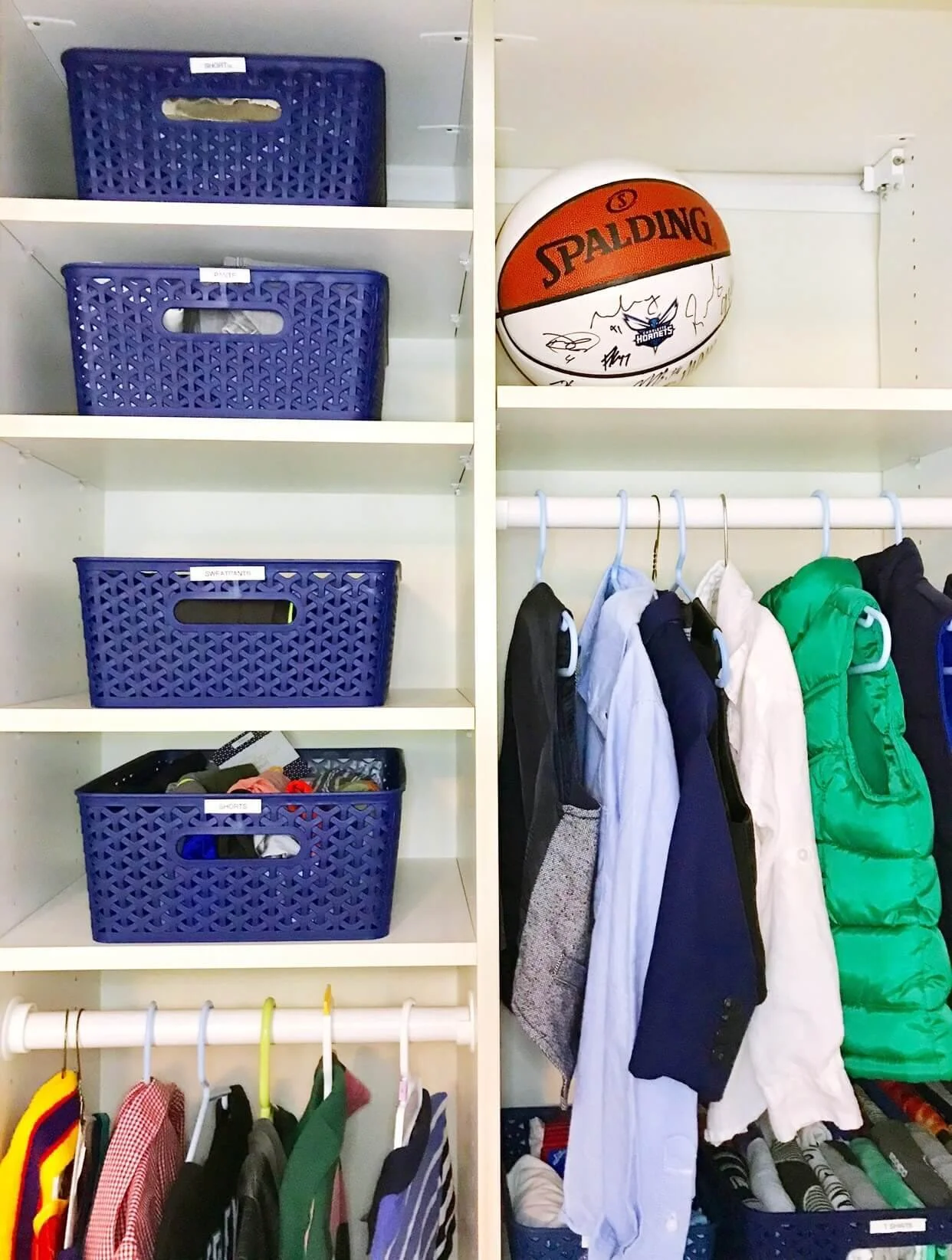  A child's closet with navy bins labeled for shorts, pants, and sweatpants, shirts and a vest on a low reachable rod, and a keepsake basketball stored on a high shelf 