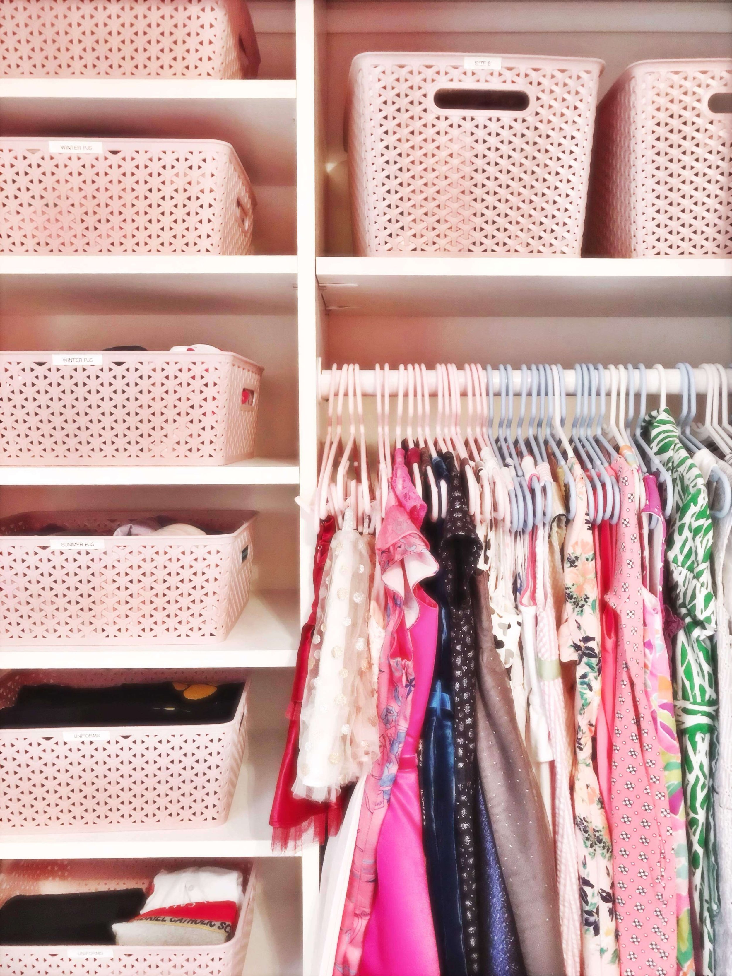  Pink woven bins labeled for winter pajamas, summer pajamas, and uniforms, stacked on shelves above a low rod of a child's hanging clothes 