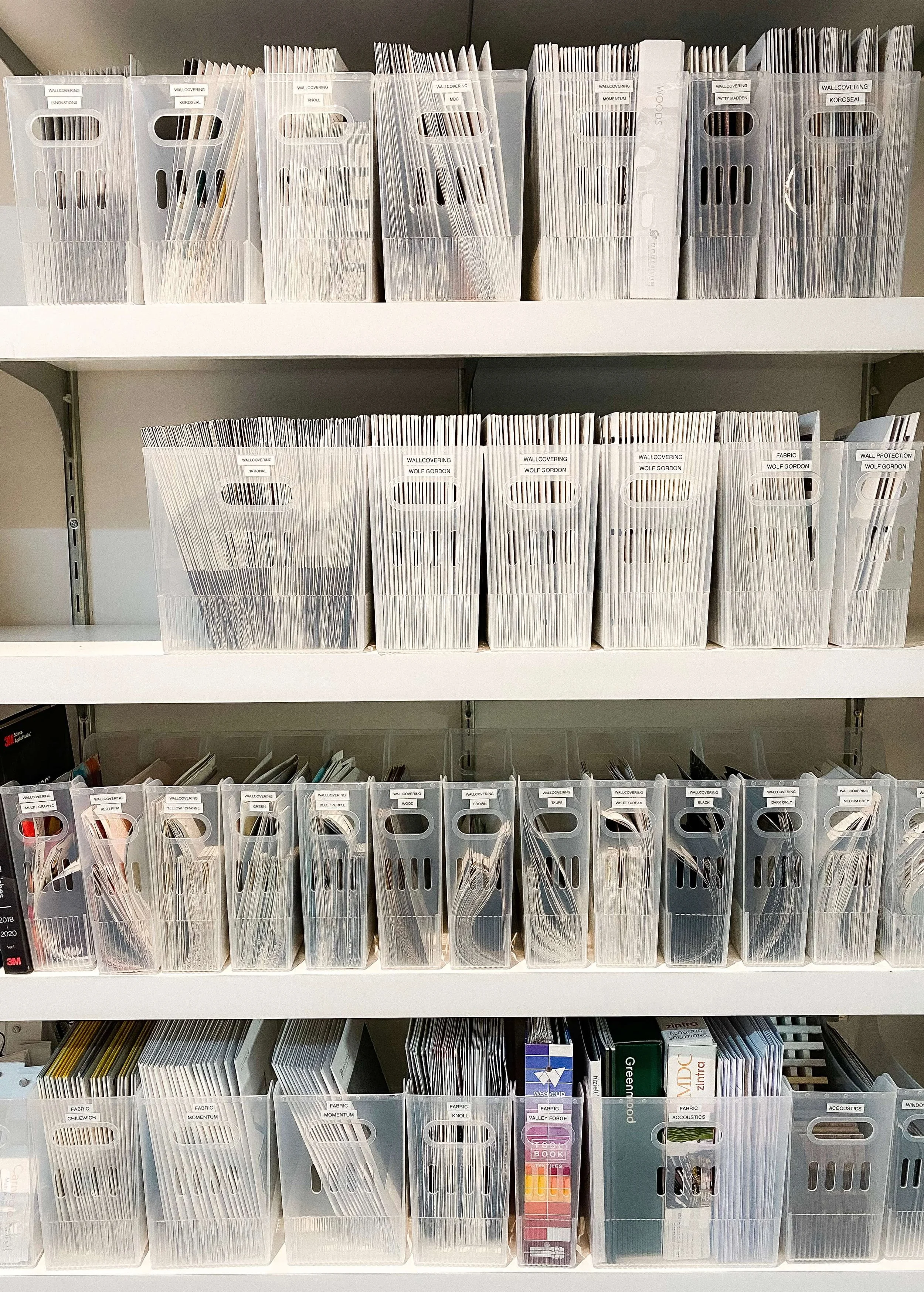 Small business organizing project for an interior design studio in Charlotte, with wallcovering, fabric, and acoustic samples sorted into clear labeled bins by vendor and color on white shelving.