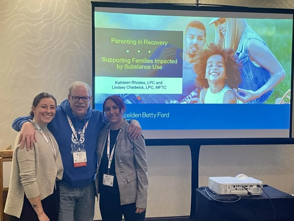 Three people smiling and standing together in front of a presentation screen at a conference, with a projector on a table nearby.