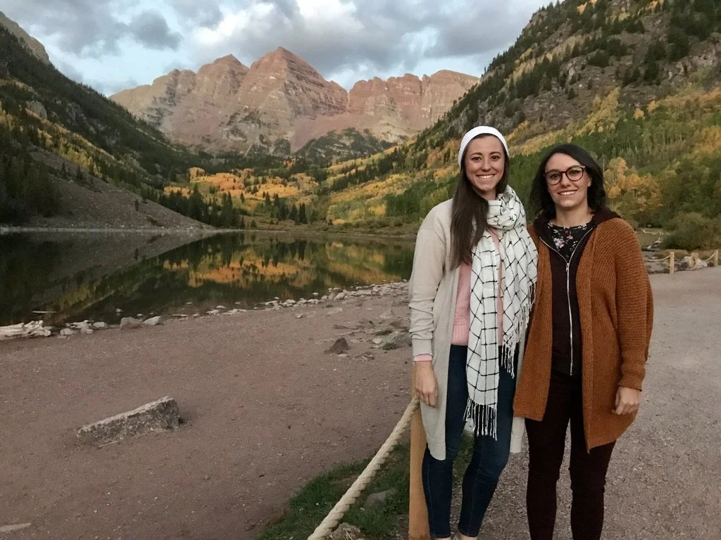 Two women smiling, standing outdoors near a lakeshore with mountains and autumn trees in the background.