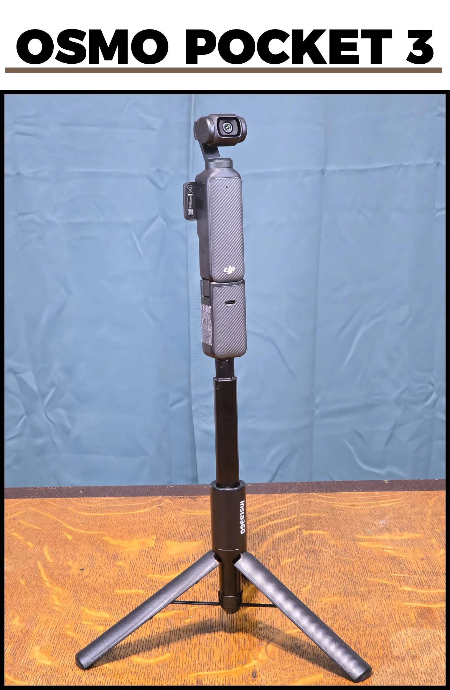 A DJI Osmo Pocket 3 handheld camera mounted on a small tripod, standing on a wooden surface against a blue backdrop.