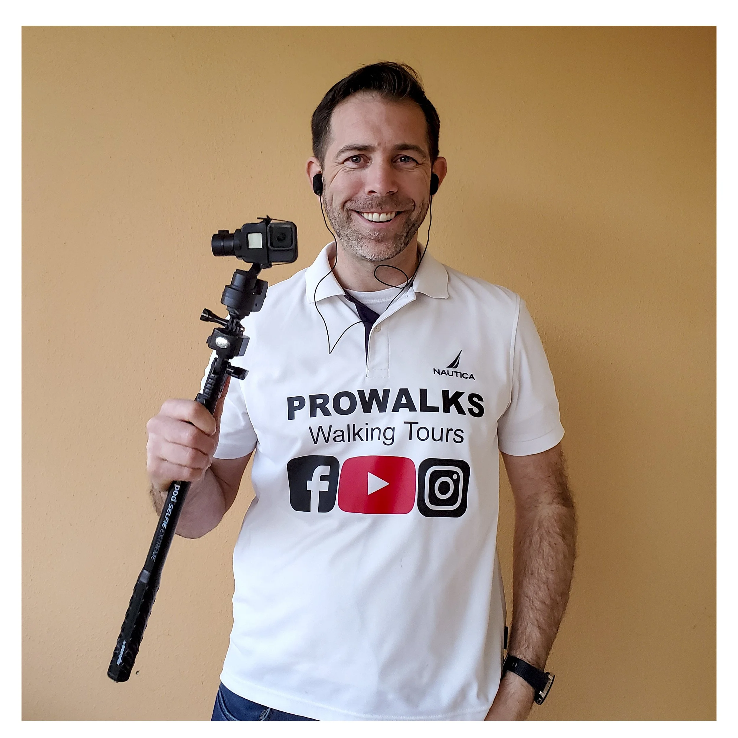 Man smiling, holding a camera mounted on a monopod, wearing a white T-shirt with social media icons and text about walking tours, standing against a plain tan wall.