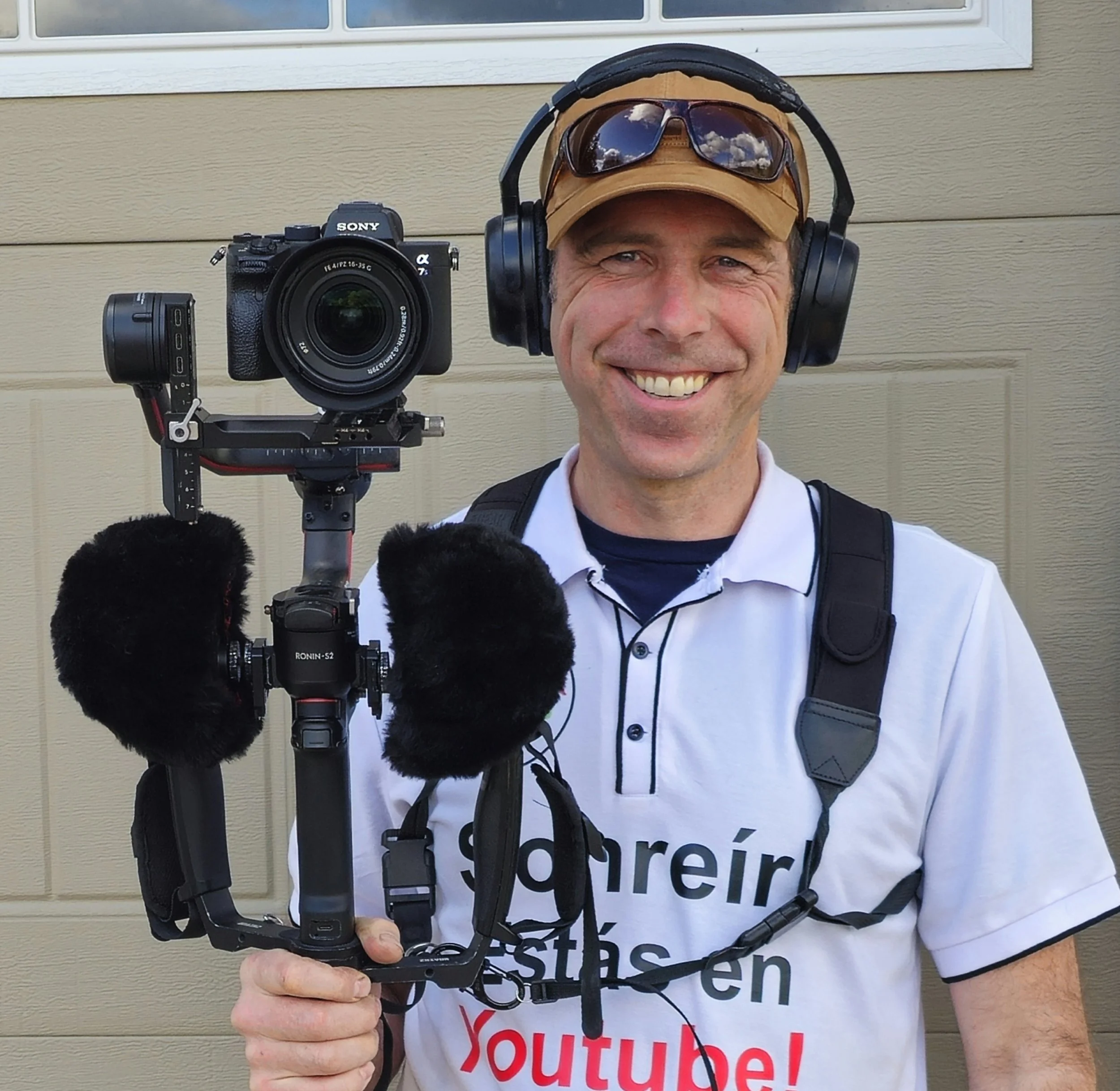 A man smiling while holding a camera stabilizer with a camera attached, wearing headphones, sunglasses, a cap, and a white shirt with partially visible text.