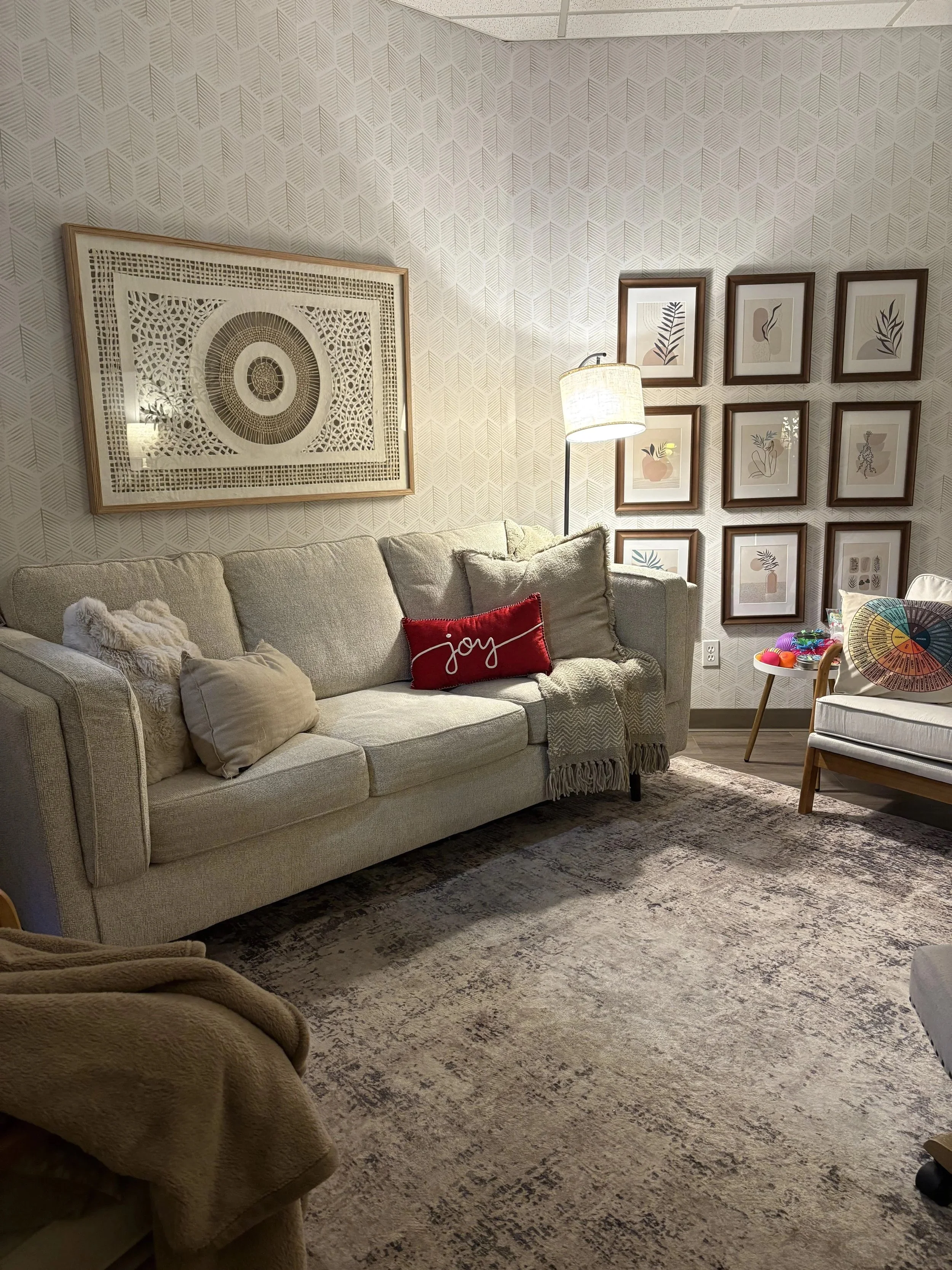 A cozy living room with a beige sofa, decorative pillows, a floor lamp, and a gallery wall of framed artwork, with a patterned wallpaper and a rug on the floor.