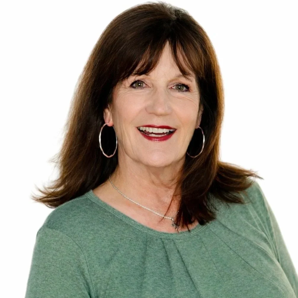 A woman with shoulder-length dark hair smiling, wearing earrings and a necklace, standing outdoors in front of green bushes.