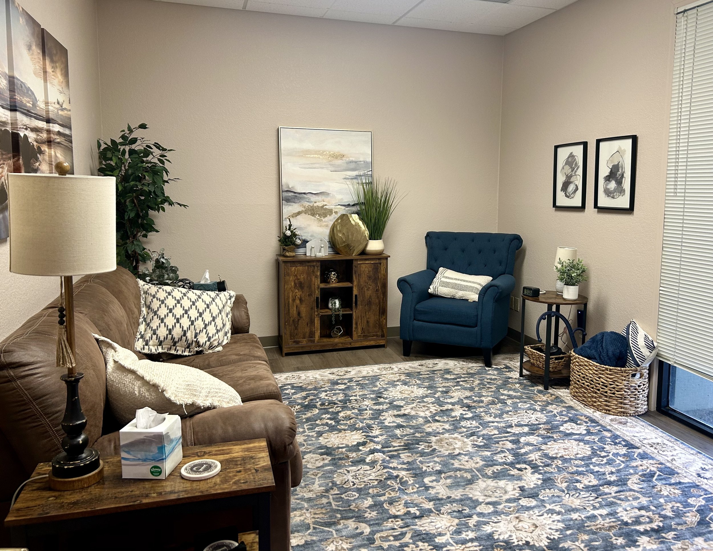 A cozy living room with a brown leather sofa on the left, a blue armchair on the right, a wooden side table with a potted plant, a large area rug with blue and beige floral patterns, and a sliding glass door with blinds on the far right. The walls ar
