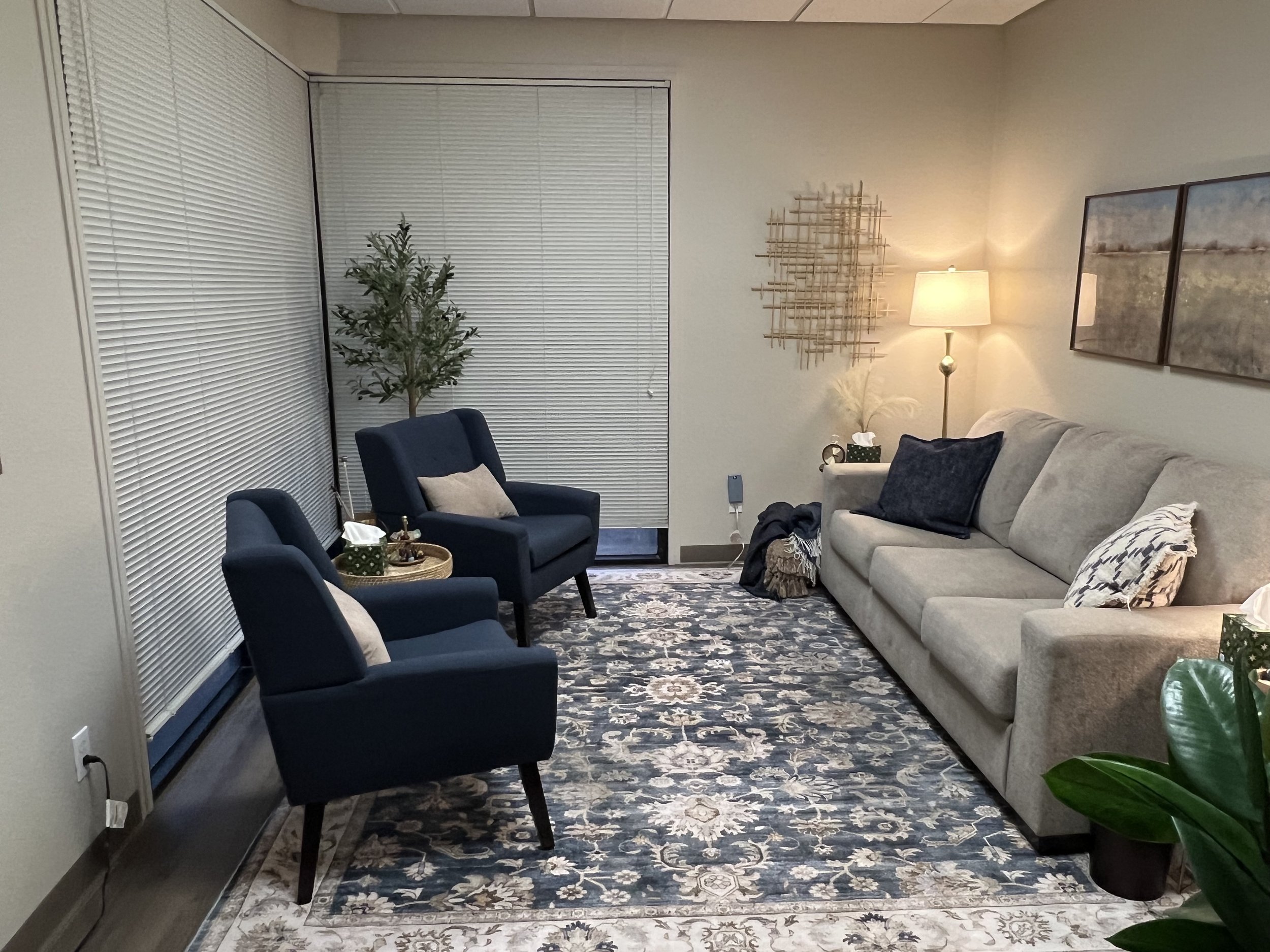 Living room with a beige couch, three decorative pillows, a floor lamp, two blue armchairs with pillows, a patterned rug, and window blinds.