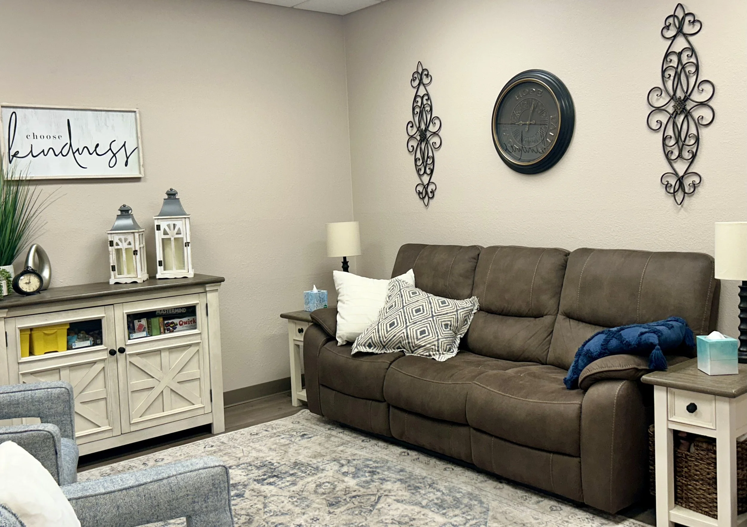 Living room with a brown leather sofa decorated with white and gray patterned pillows, two white end tables with lamps, a wall clock and wall art, and a cream-colored cabinet with lanterns and decorative items on top.