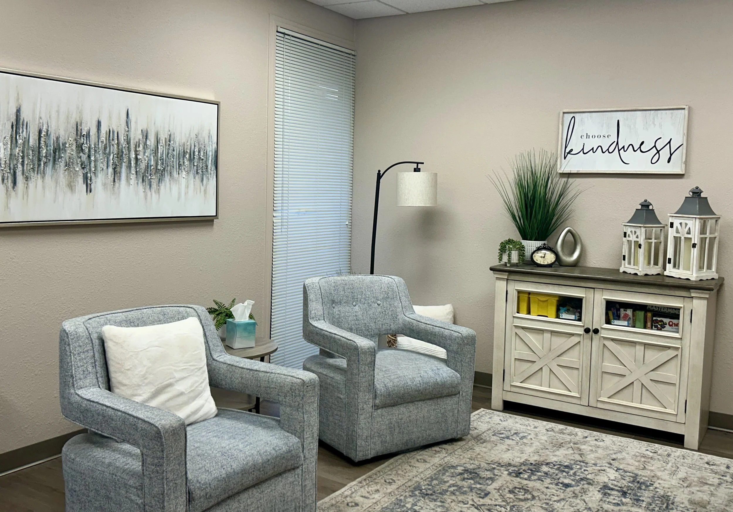A cozy waiting room with two gray armchairs, a side table with a tissue box, a white cabinet with books and decorative items, a large abstract painting, and a wall sign saying 'choose kindness'.