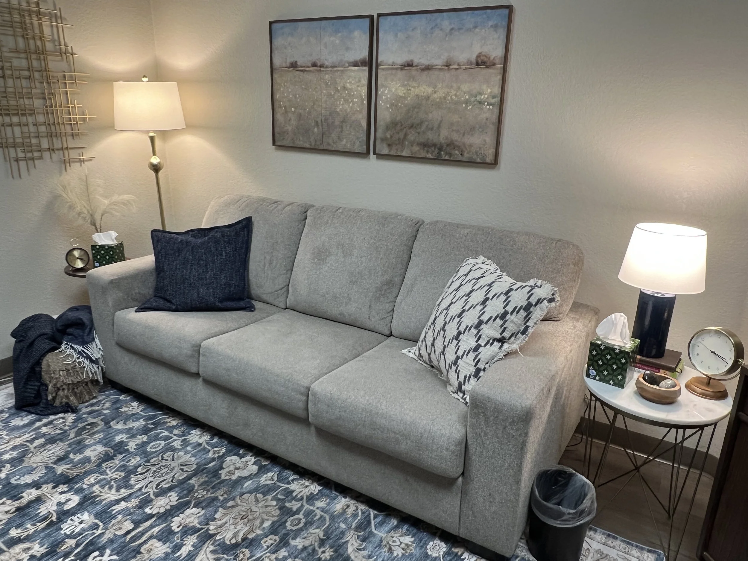 A living room with a light beige sofa adorned with two decorative pillows, a white side table with a lamp, tissue box, clock, and other small items, a patterned blue and white rug, and wall art including a landscape painting and a gold geometric wall