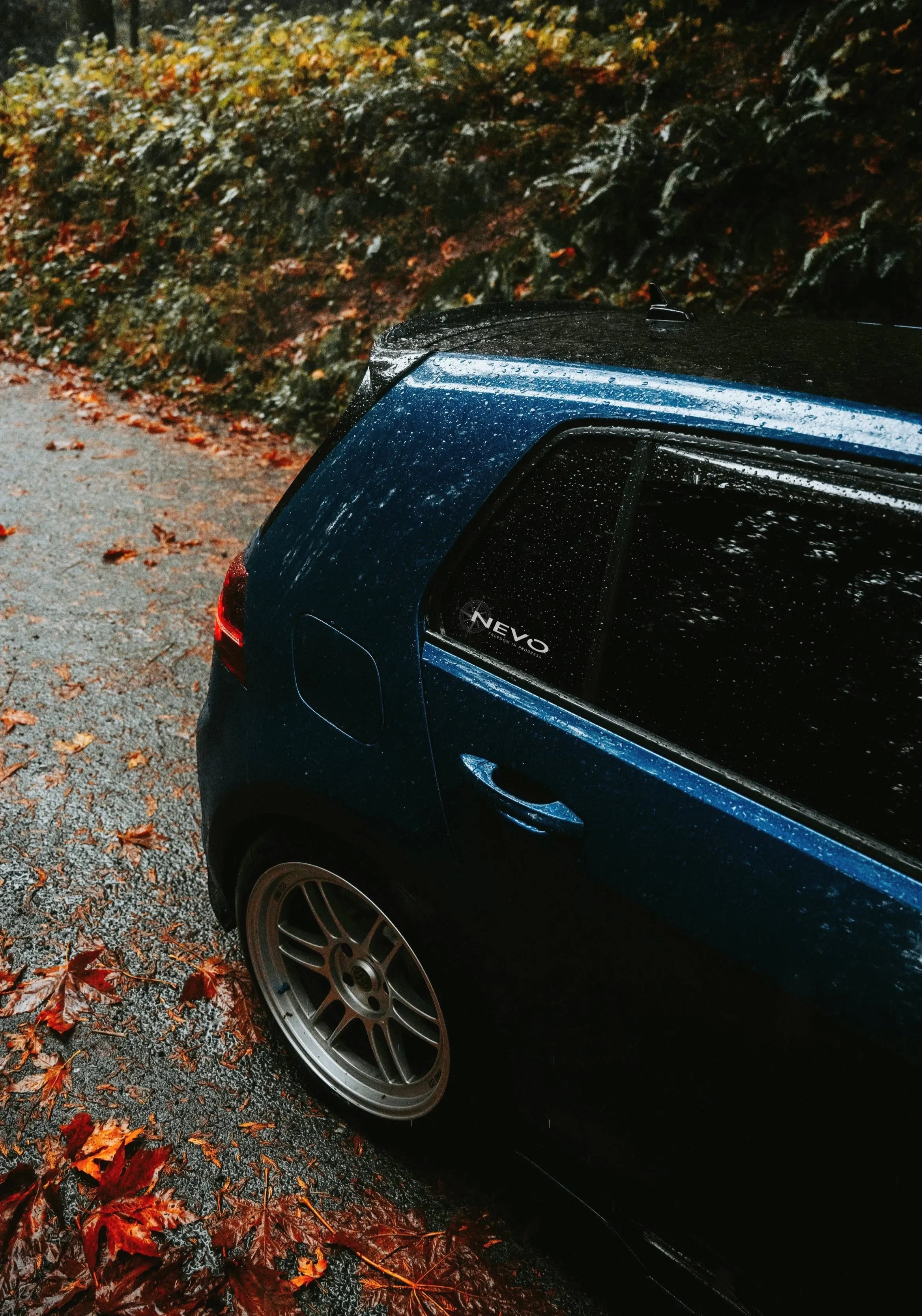 Blue NEVO Driving Academy car on a rainy forest road surrounded by fallen leaves, capturing the spirit of fall driving safety and exploration.