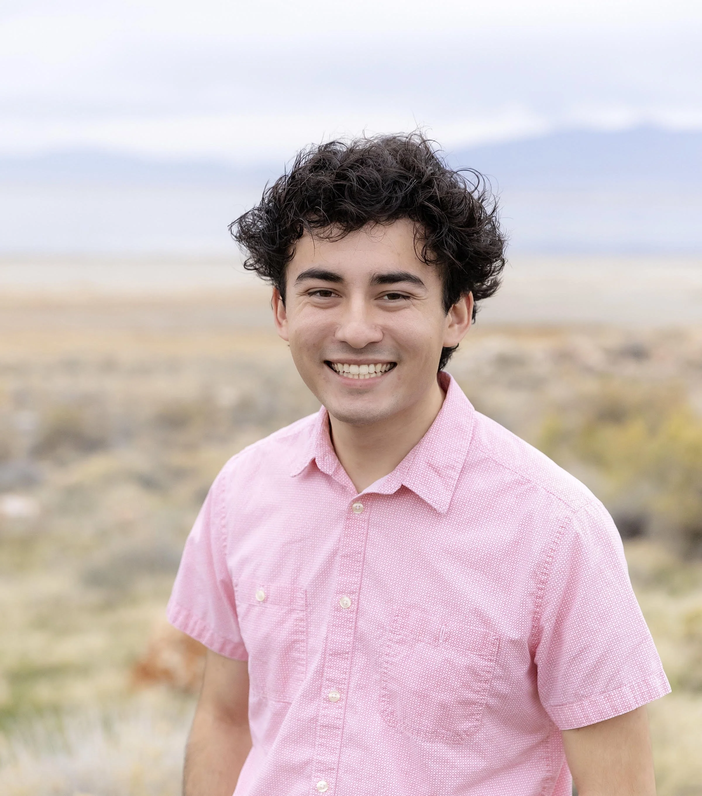 A young man with curly dark hair, wearing a pink button-up shirt, smiling outdoors with a blurred landscape and overcast sky in the background.