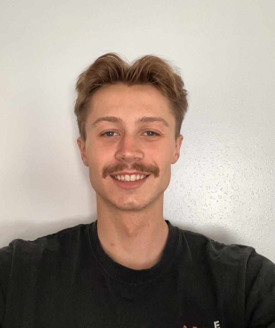 A young man with short, light brown hair, a mustache, and a light complexion smiling at the camera, wearing a black T-shirt, standing against a plain white wall.