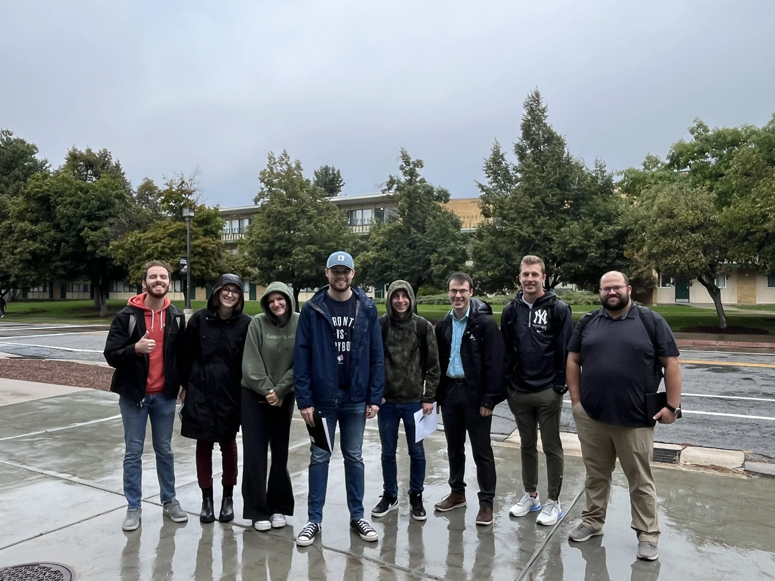 Group of eight people standing outdoors on a sidewalk, smiling and posing for the photo, with trees and a building in the background on a rainy day.