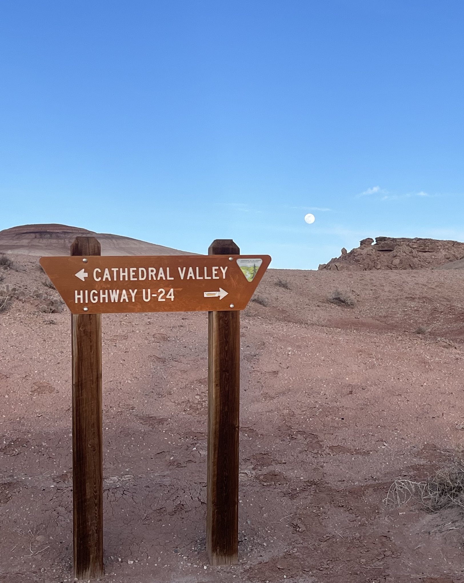 A wooden trail sign in a desert landscape with a blue sky and visible moon; arrows pointing left and right to Cathedral Valley and Highway U-24.