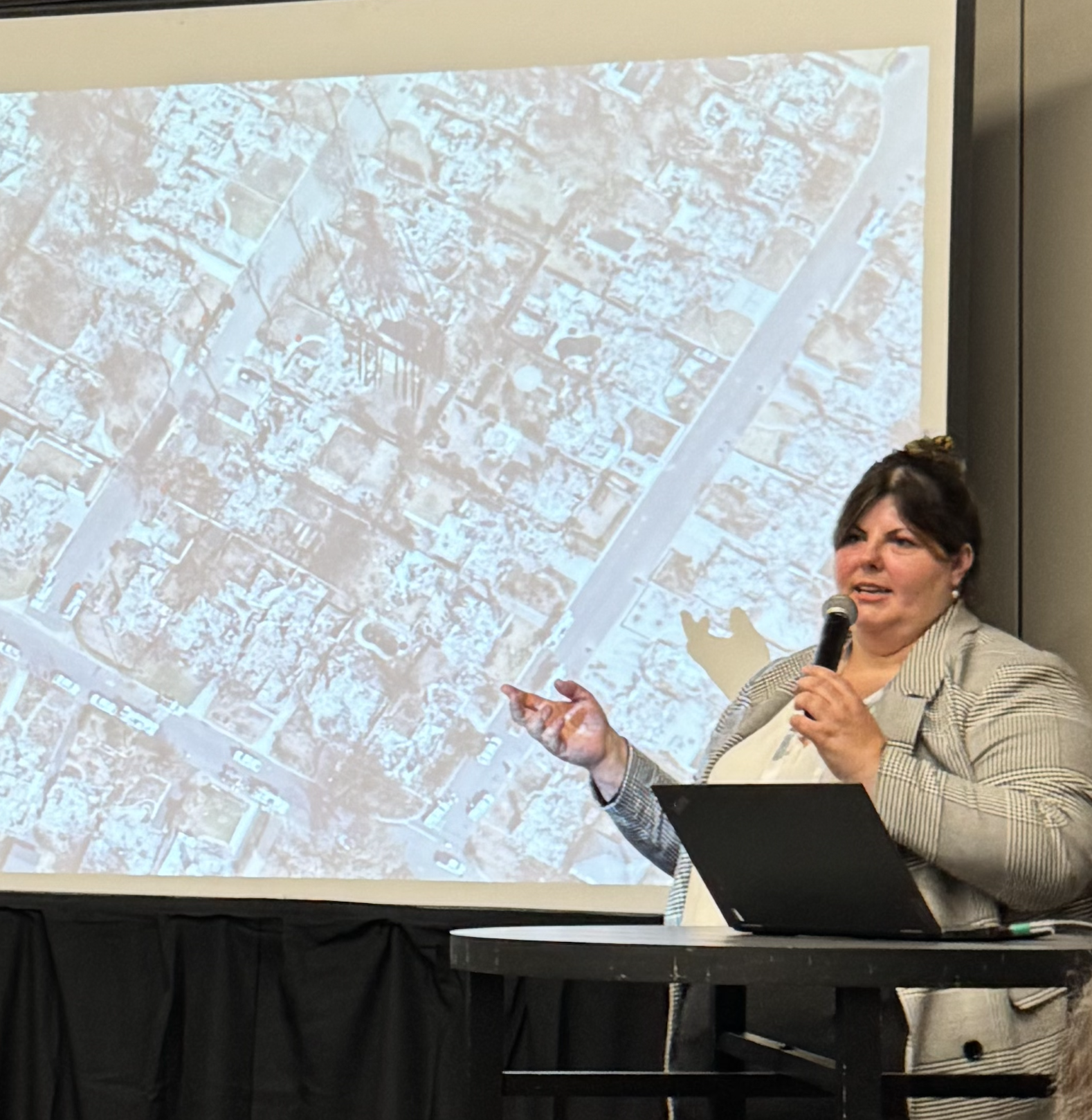 A woman giving a presentation using a microphone, standing in front of a large screen displaying a map or aerial view of a city.