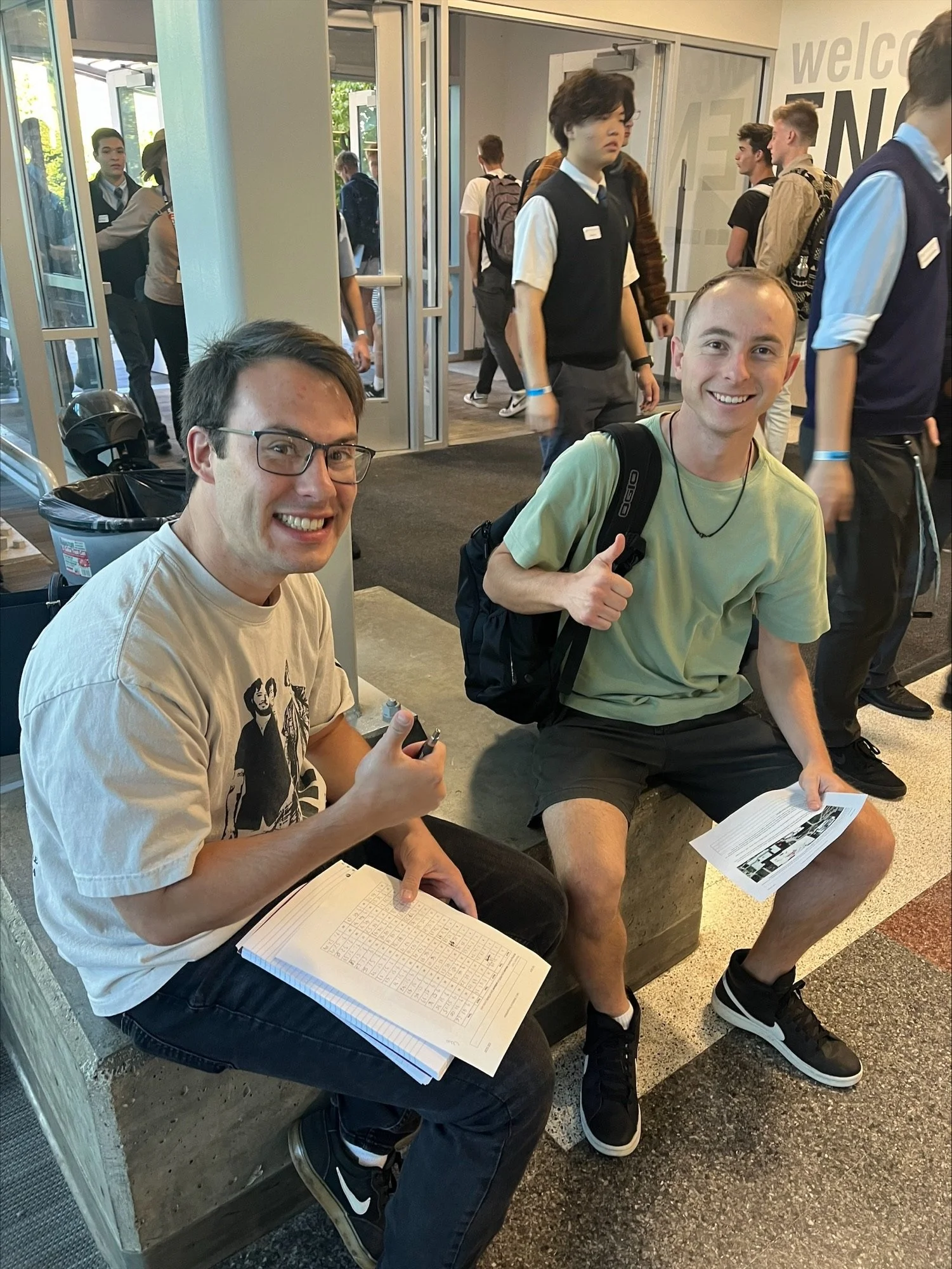 Two young men sitting on a bench at school. They are smiling and holding papers, with one giving a thumbs-up. There are students walking in the background near an open door entrance.