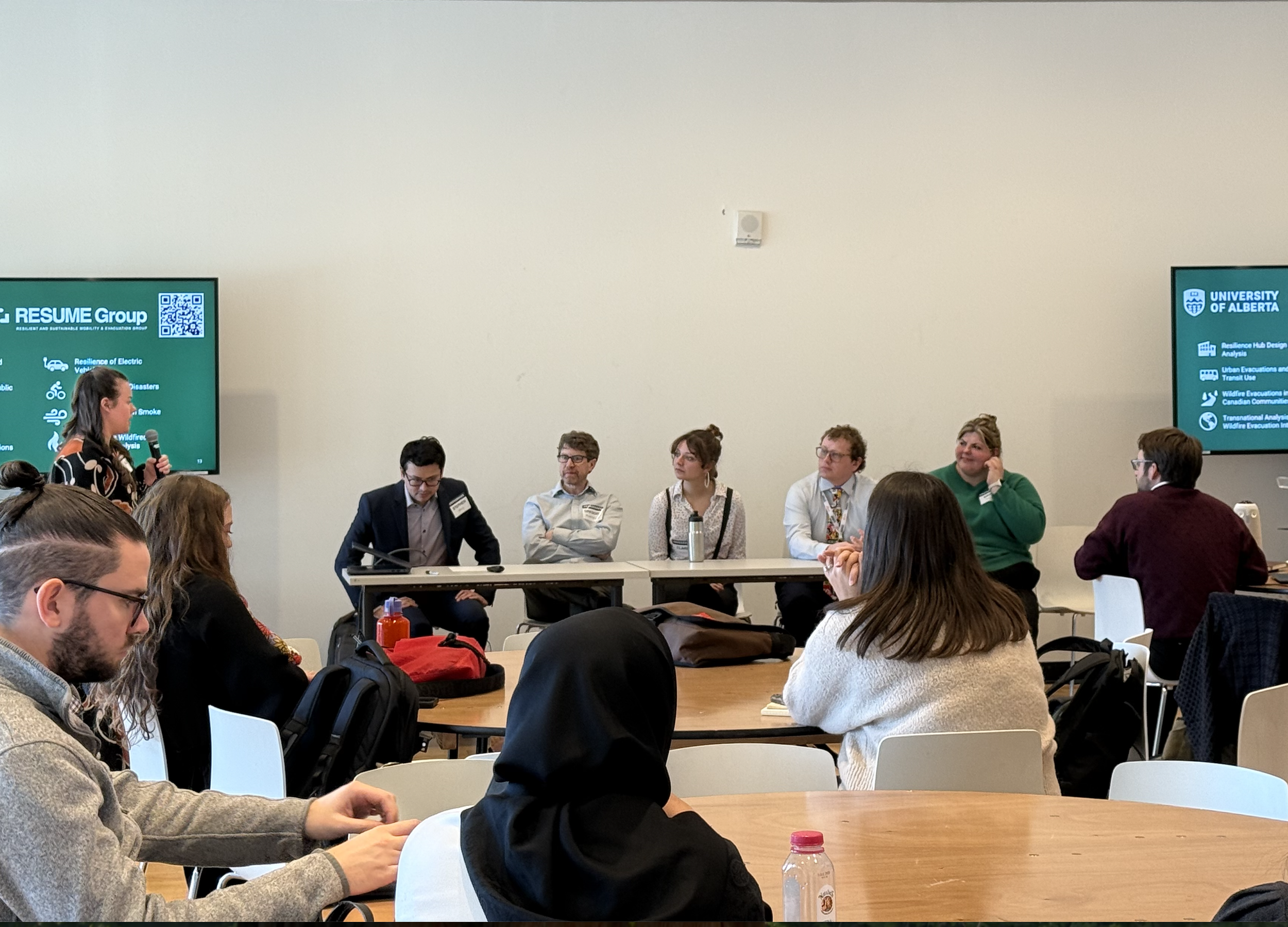 A woman is speaking at a panel discussion in a conference room, with five panelists seated at a long table. Audience members are seated at round tables, listening attentively. Two screens display presentation slides related to the University of Alberta.