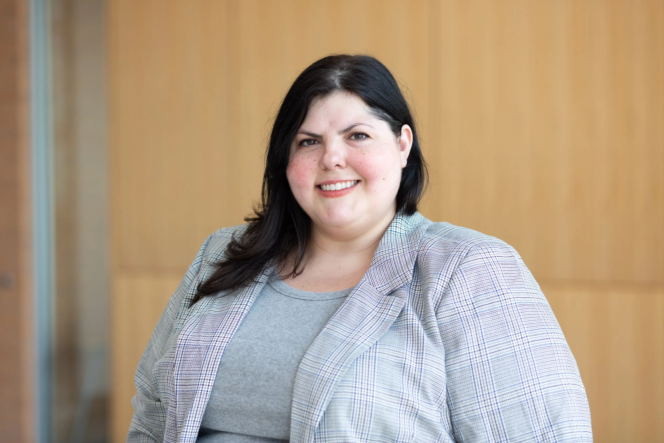 Portrait of a smiling woman with black hair wearing a plaid blazer over a gray top, standing in front of a wooden paneled wall.