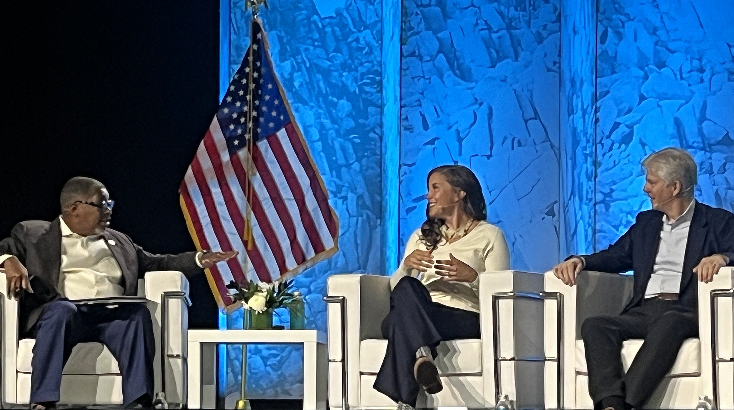 Three individuals sitting on stage in a panel discussion, with an American flag and a blue background. The person on the left is an older man with glasses, wearing a dark suit and yellow shirt. The woman in the middle is smiling, with long dark hair, wearing a light-colored top. The man on the right is also smiling, with gray hair, wearing a dark blazer and white shirt. There is a small table with a flower arrangement in front of them.