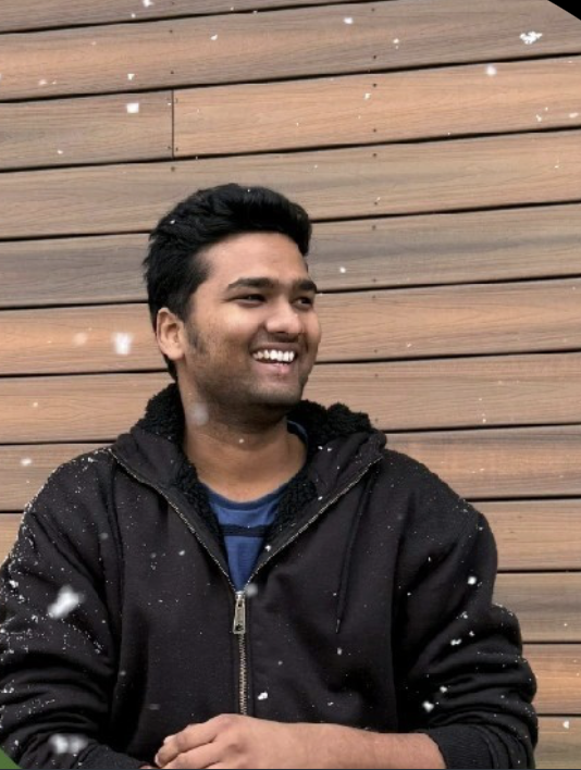 A young man with dark hair, smiling, wearing a black hoodie, standing against a wooden wall background with falling snow.