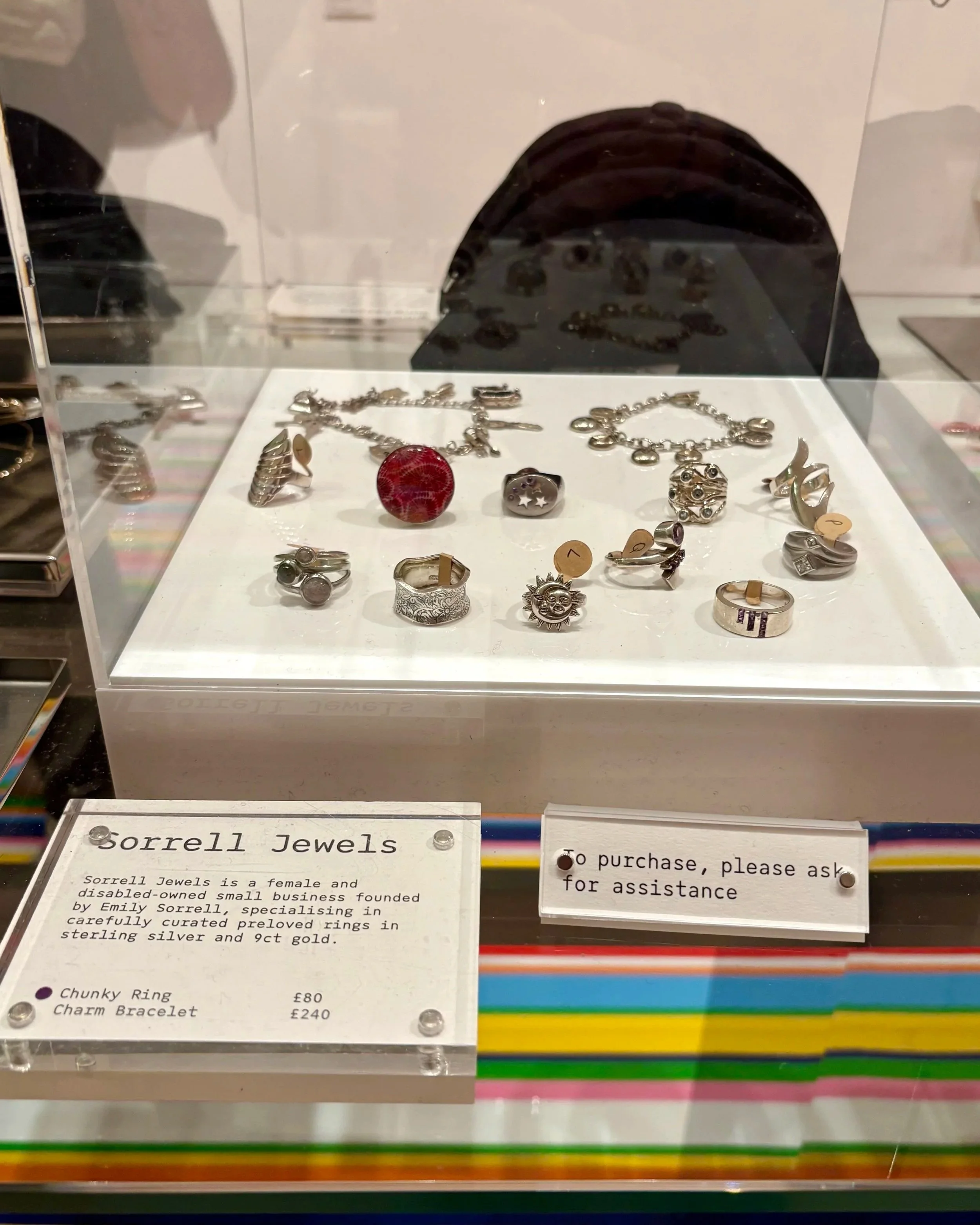 A glass display case in the Tate Britain shop filled with vintage sterling silver rings and two charm bracelets on a white stand, with a Sorrell Jewels sign beside it, set on a colourful striped counter.