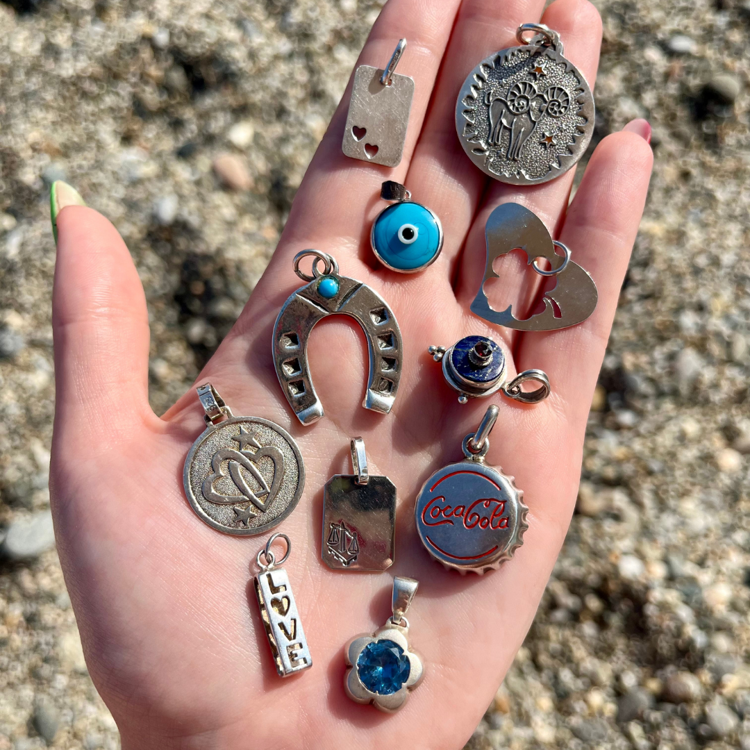 A hand holding a curated selection of vintage silver charms, including lucky symbols and engraved pendants, photographed outdoors on a pebbled surface during a jewellery sourcing trip.