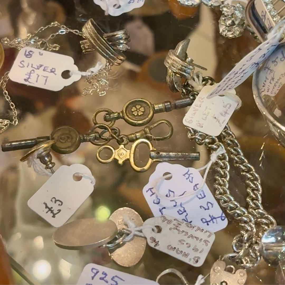 Close-up of vintage silver jewellery and chains displayed inside a glass cabinet, each piece labelled with handwritten price tags at an antiques fair.