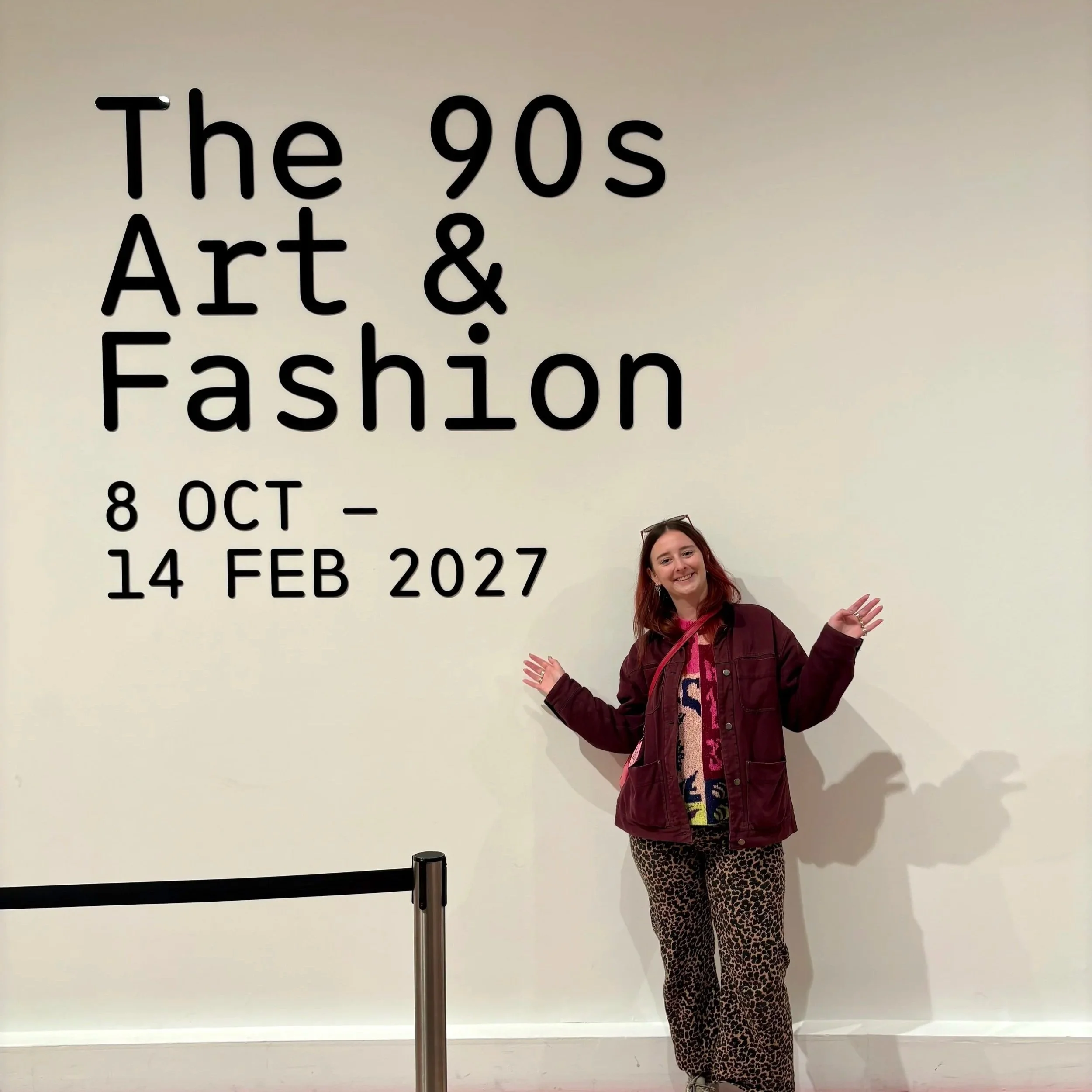Me smiling with my arms out in front of the wall sign for The 90s: Art and Fashion exhibition at Tate Britain, showing the dates 8 October to 14 February 2027.