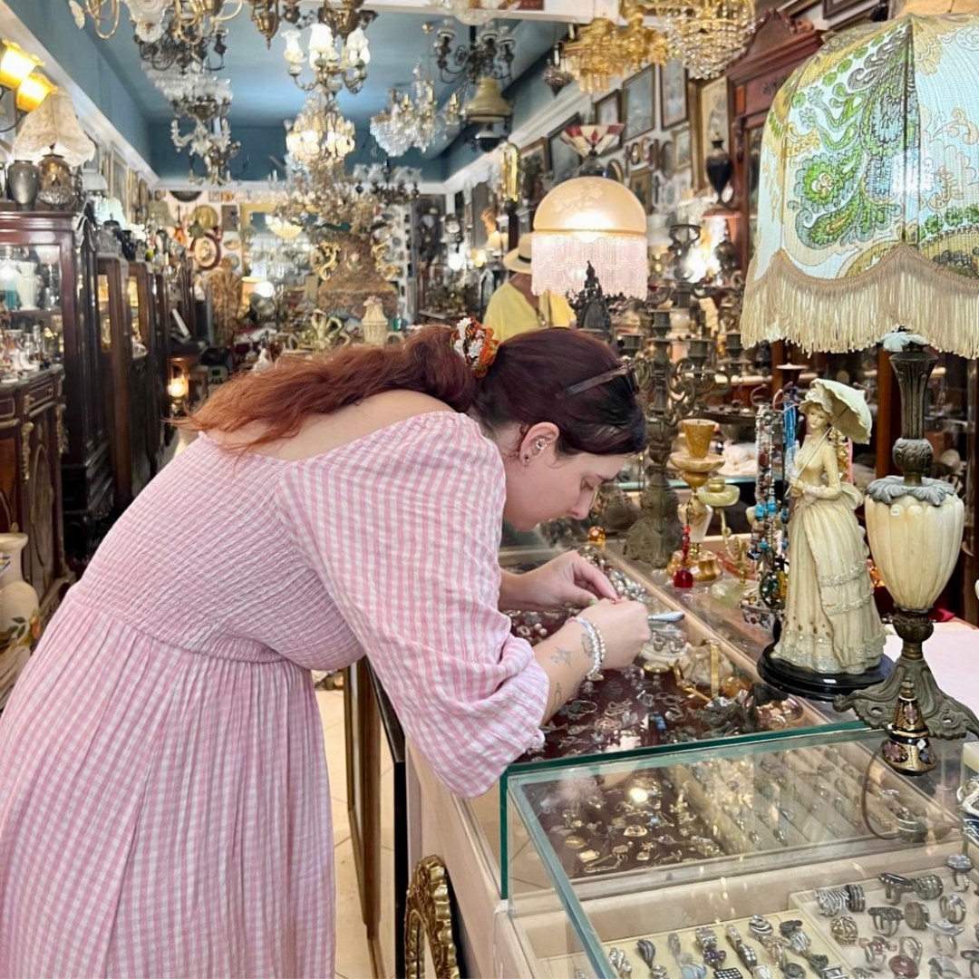A woman browsing vintage jewellery inside a characterful antique shop filled with chandeliers, glass cabinets, and curated treasures during a sourcing trip.