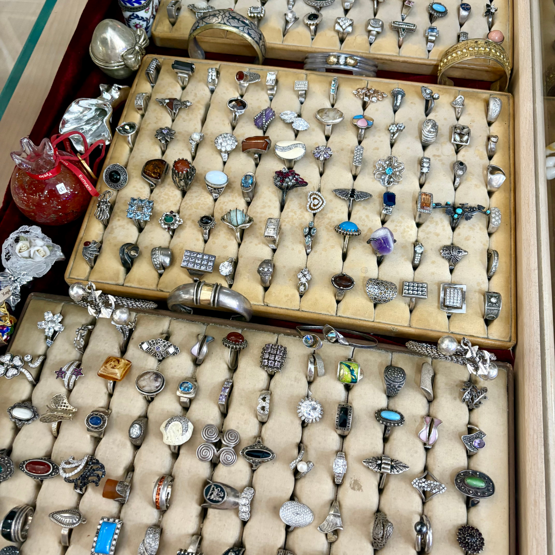 Rows of vintage and antique rings displayed in velvet-lined trays, featuring a mix of silver and gemstone designs at a jewellery market or auction.