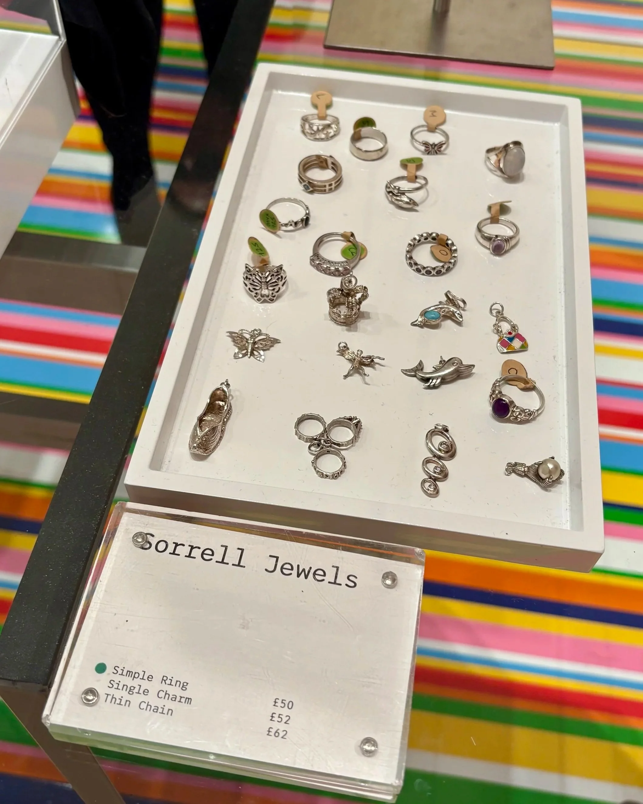 A white tray of vintage sterling silver rings and charms in the Tate Britain shop, including dolphin, butterfly, crown and shoe charms and rings set with purple stones, beside a Sorrell Jewels price sign on a colourful striped counter.
