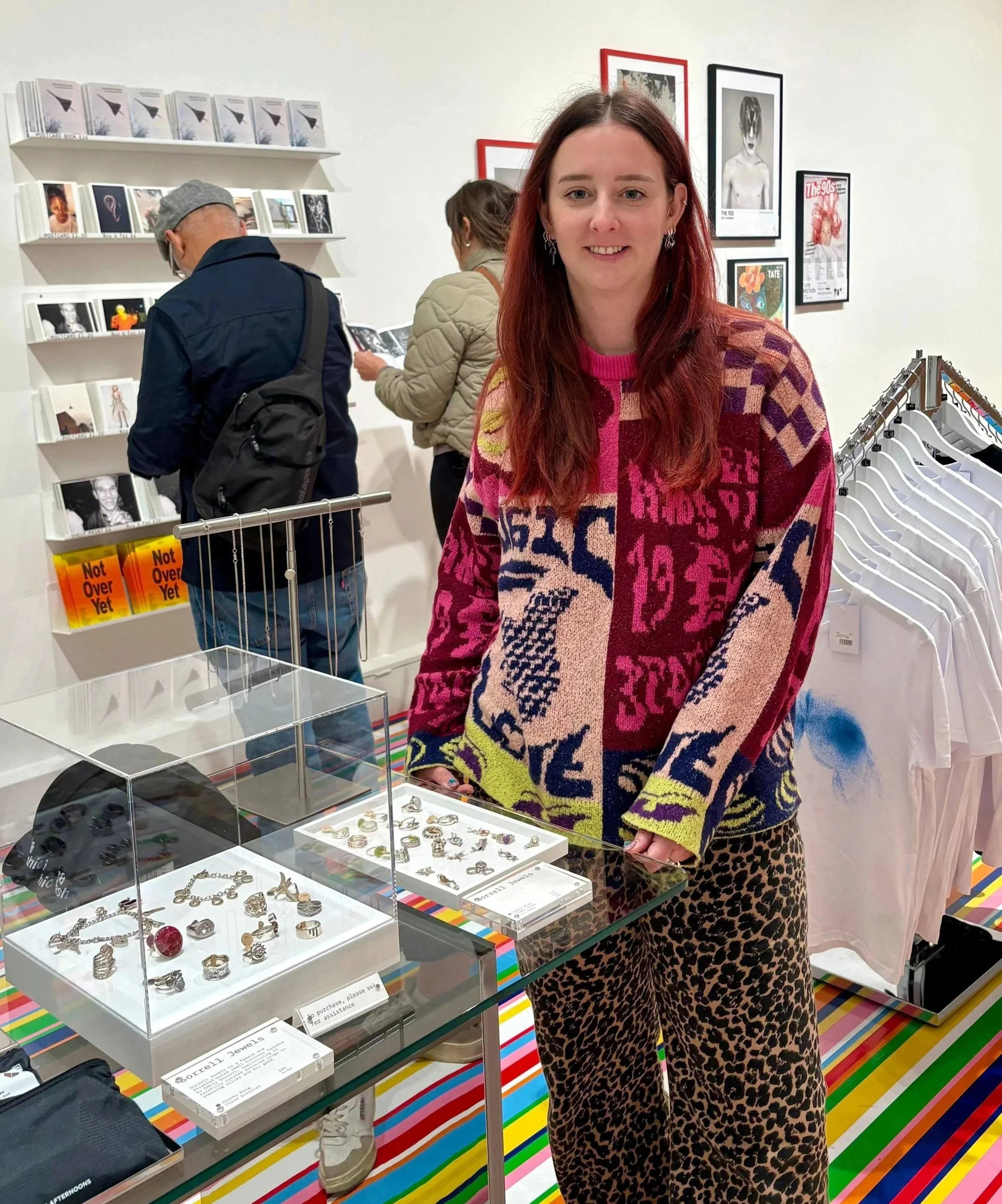 Me standing beside the Sorrell Jewels display in the Tate Britain shop, with vintage silver rings and charm bracelets in glass cases on a colourful striped counter, and shoppers browsing prints and postcards in the background.