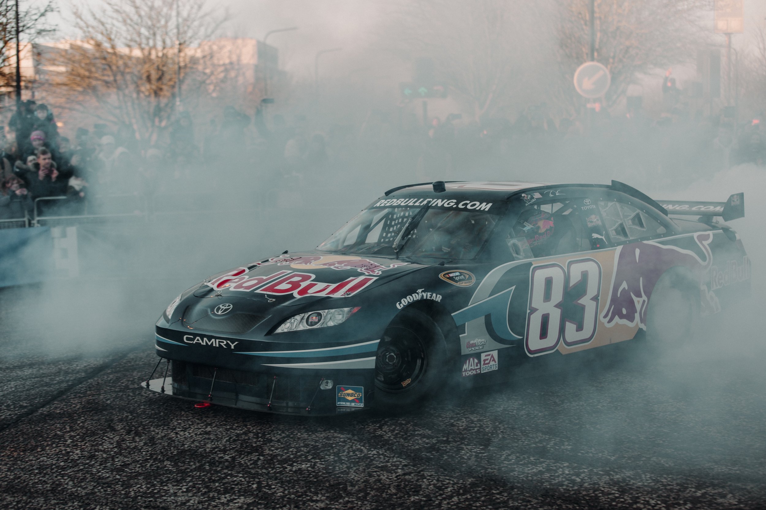 A black race car with Red Bull branding, number 83, performing a burnout with smoke in the air, spectators watching in the background.