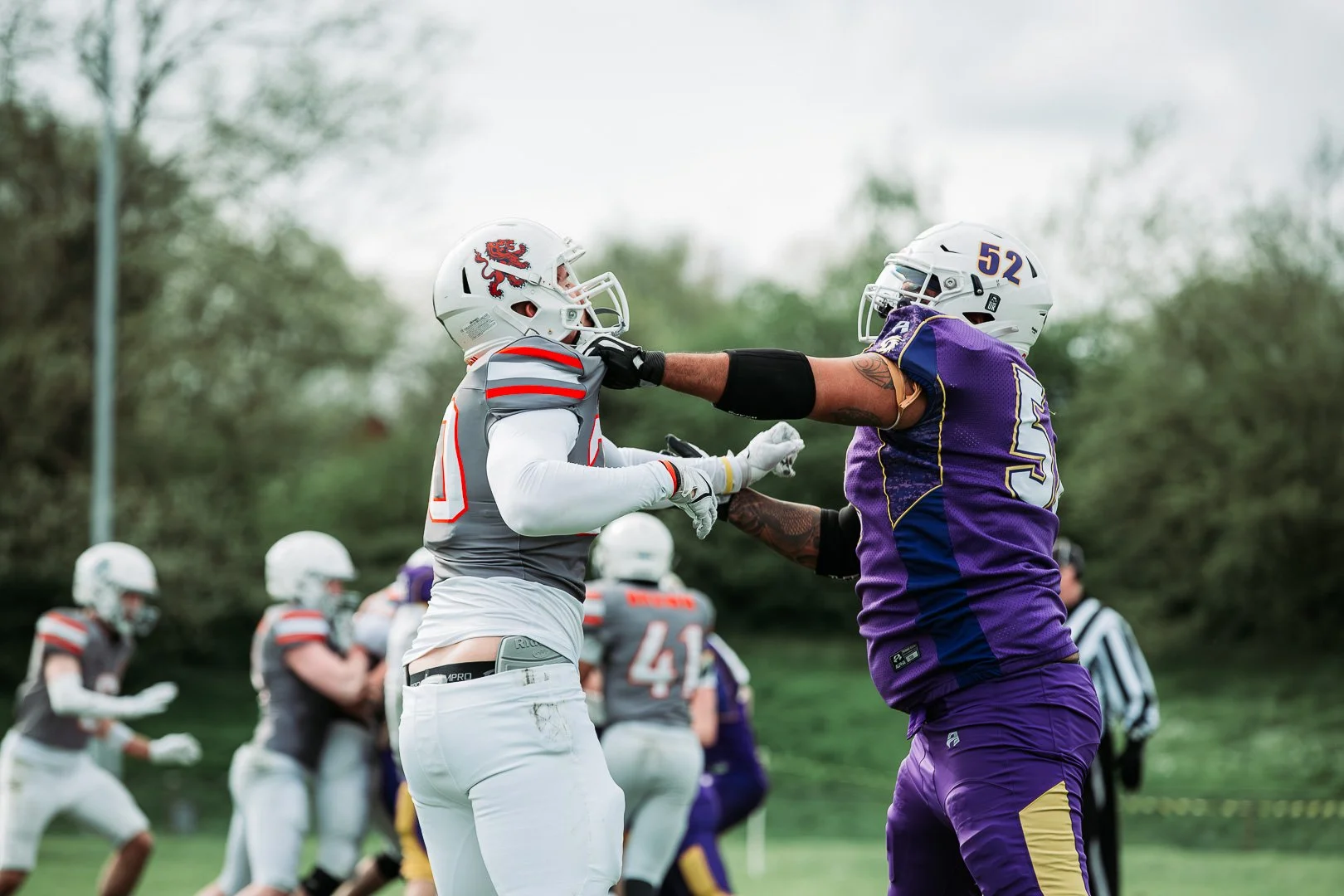 Two football players, one in a gray and red uniform and the other in a purple and yellow uniform, face off on a football field during a game or practice, with additional players and an official in the background.